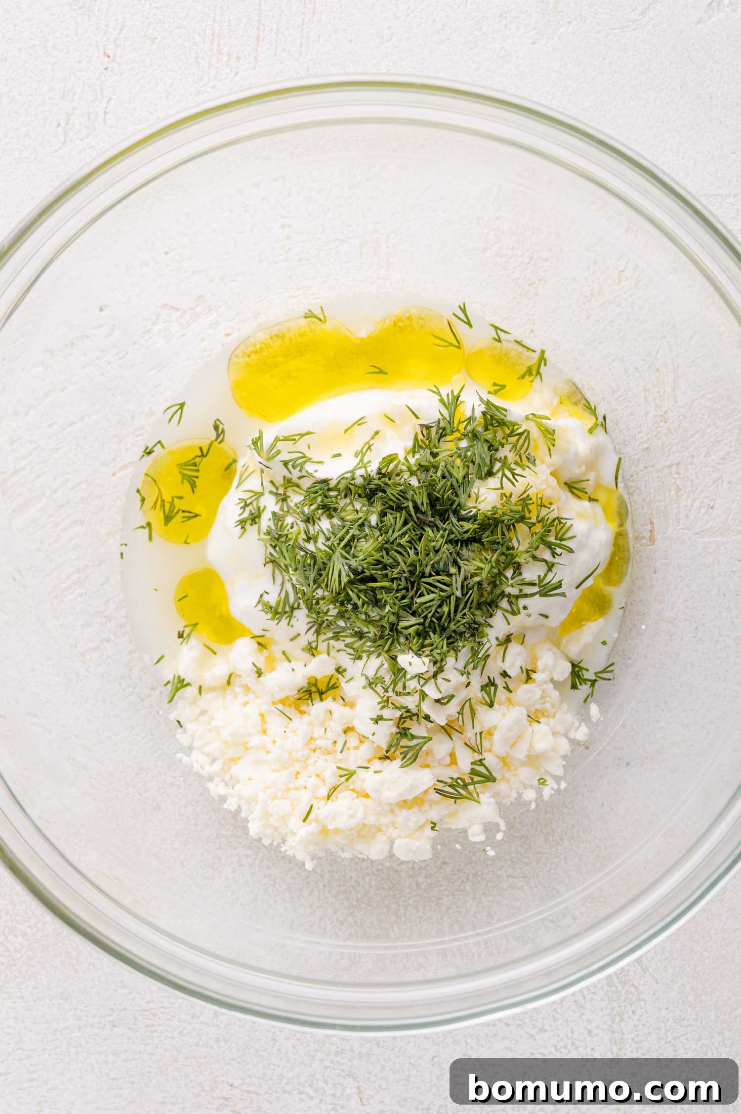 A clear glass bowl on a light surface holds a blend of creamy plain yogurt, tangy feta cheese, fresh chopped dill, olive oil, and a splash of lemon juice, ready to be whisked into a delectable sauce.