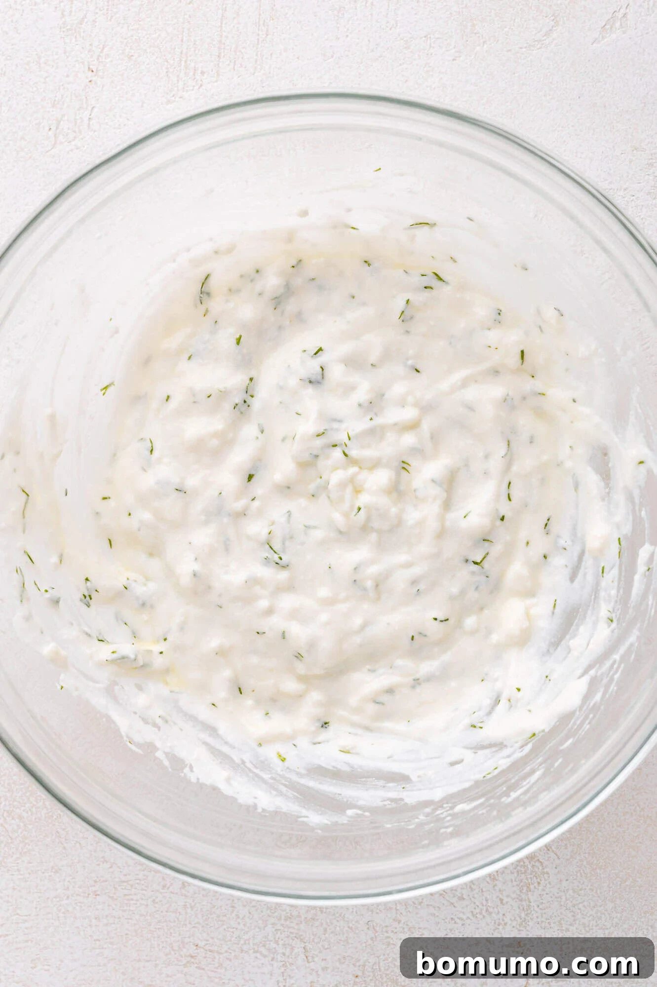 A clear glass bowl filled with a luxurious, creamy white mixture, visibly flecked with vibrant green herbs, resting invitingly on a pristine light surface. The sauce has a smooth, enticing texture.