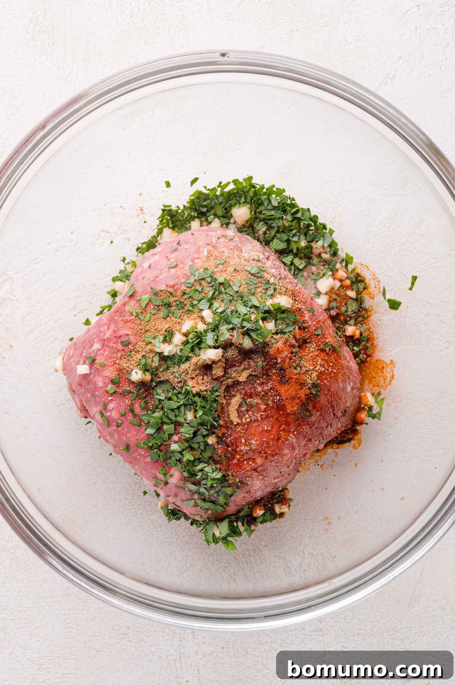 A raw mound of ground lamb rests in a clear glass bowl, generously sprinkled with an array of vibrant chopped herbs, finely minced garlic, and a rich blend of various spices, all presented on a clean white surface, ready for mixing.