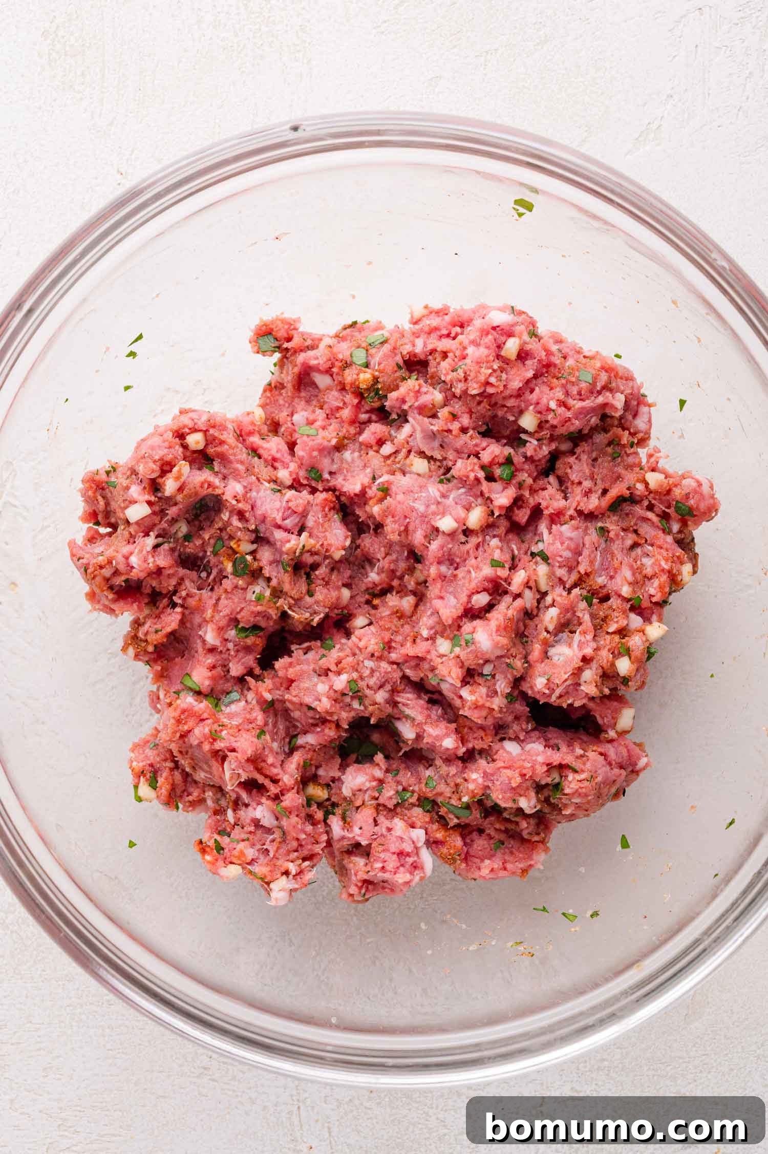 A clear glass bowl showcasing a perfectly seasoned raw ground lamb mixture, artfully combined with fresh herbs and an aromatic blend of spices, all resting on a bright, light-colored surface, indicative of readiness for patty formation.