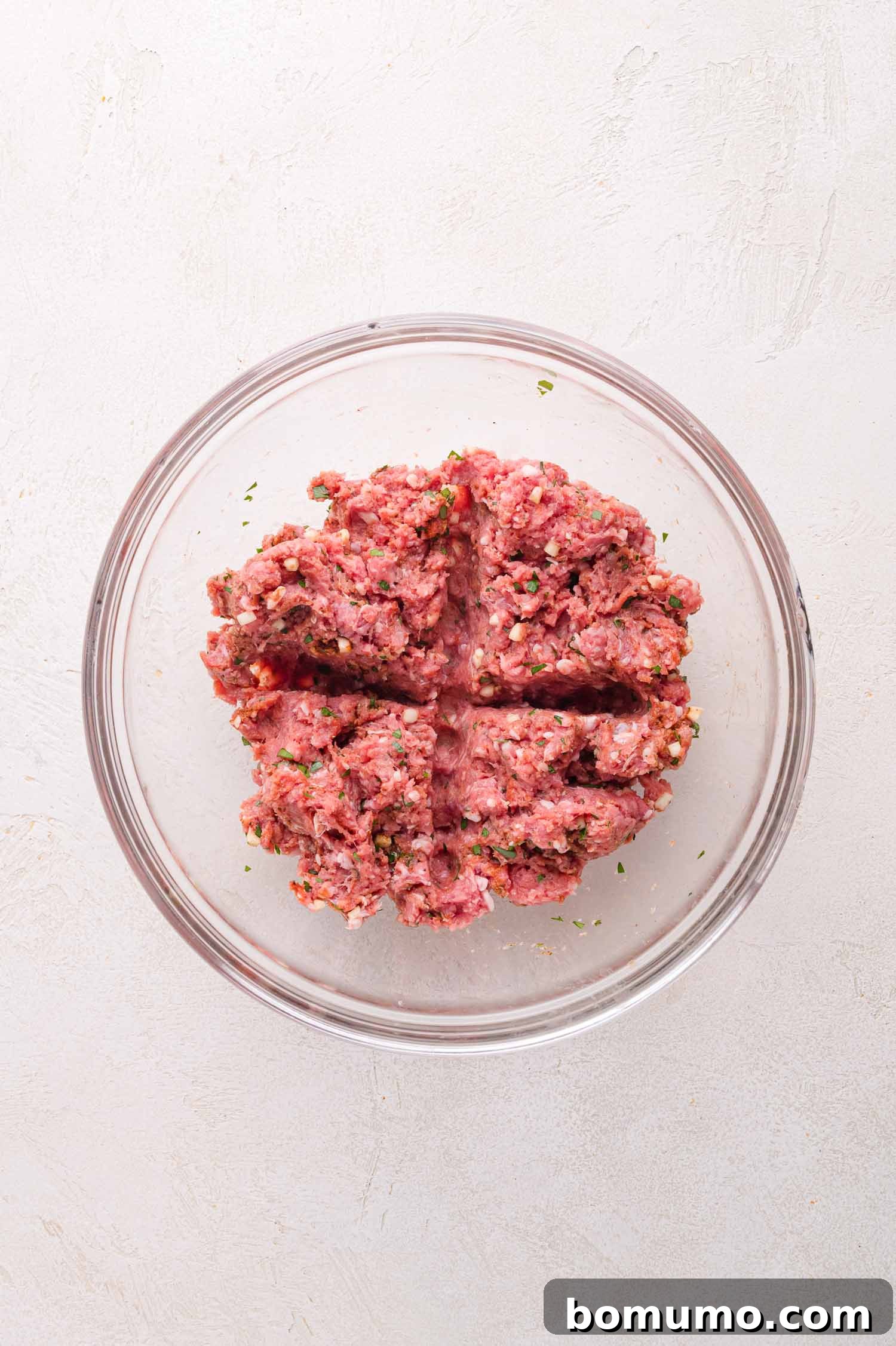 A clear glass bowl containing a perfectly seasoned raw ground lamb mixture, precisely divided into four equal sections, signifying readiness for shaping into patties. The arrangement is on a bright, light-colored surface.