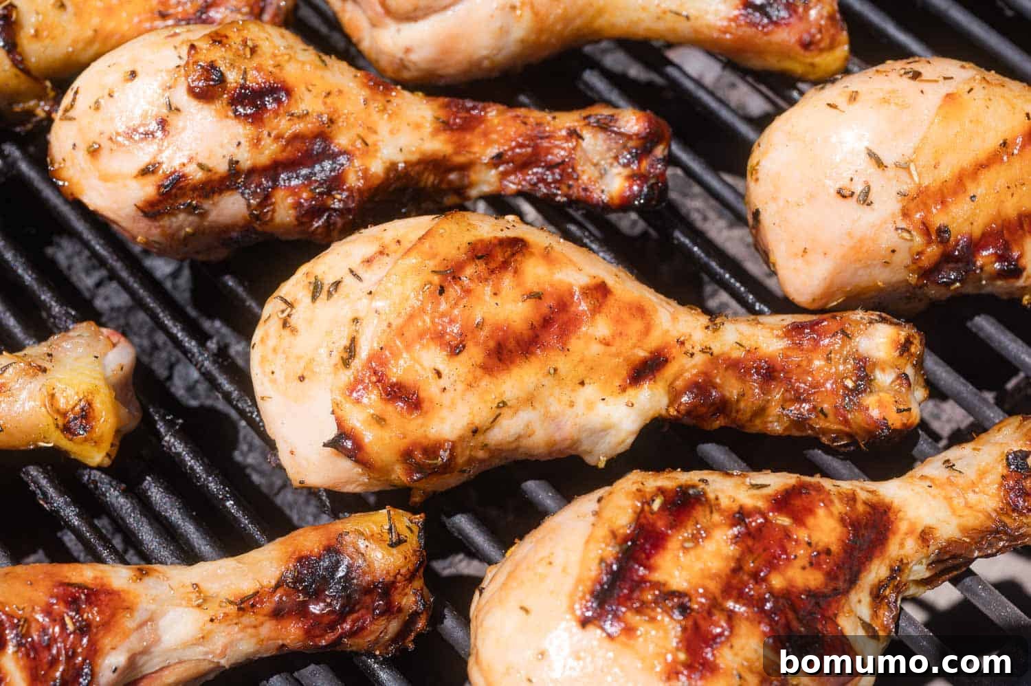 Several seasoned chicken drumsticks cooking on a grill with visible char marks and smoke rising.