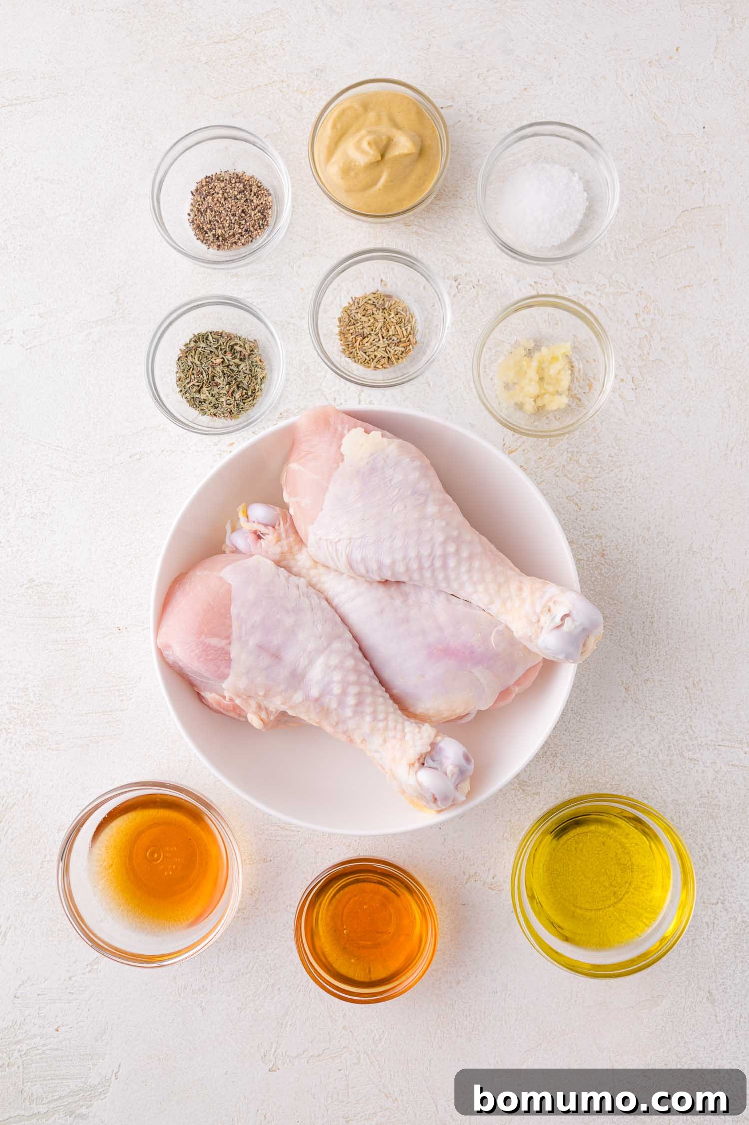 A bowl of raw chicken drumsticks surrounded by small bowls of various spices and seasonings, arranged on a white surface, preparing for marinade.