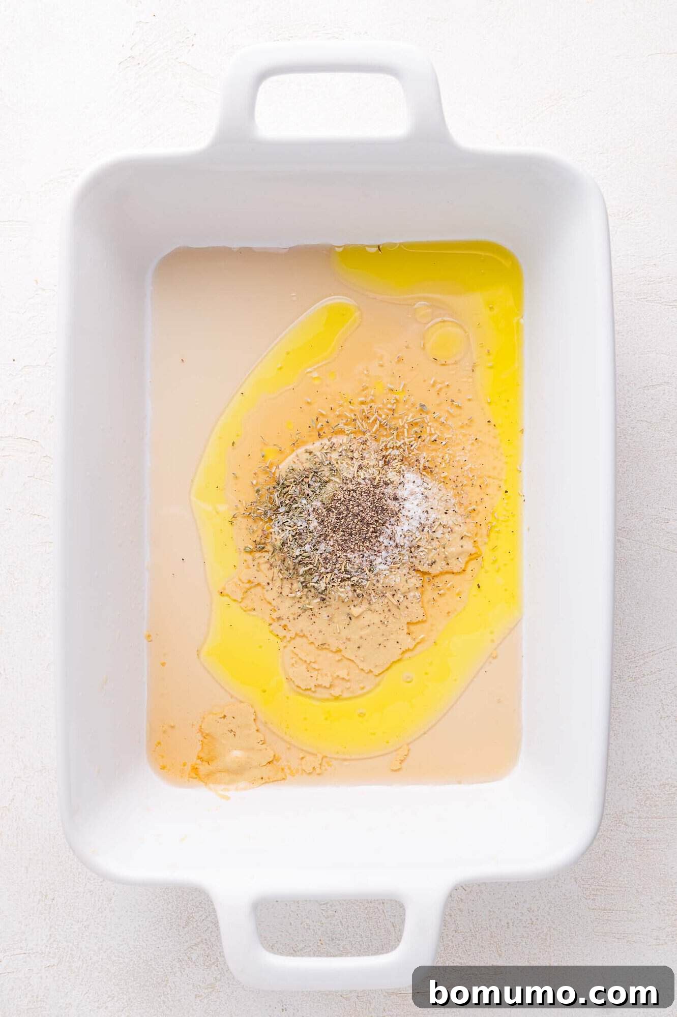 A white baking dish on a white surface, containing the initial marinade ingredients before mixing.
