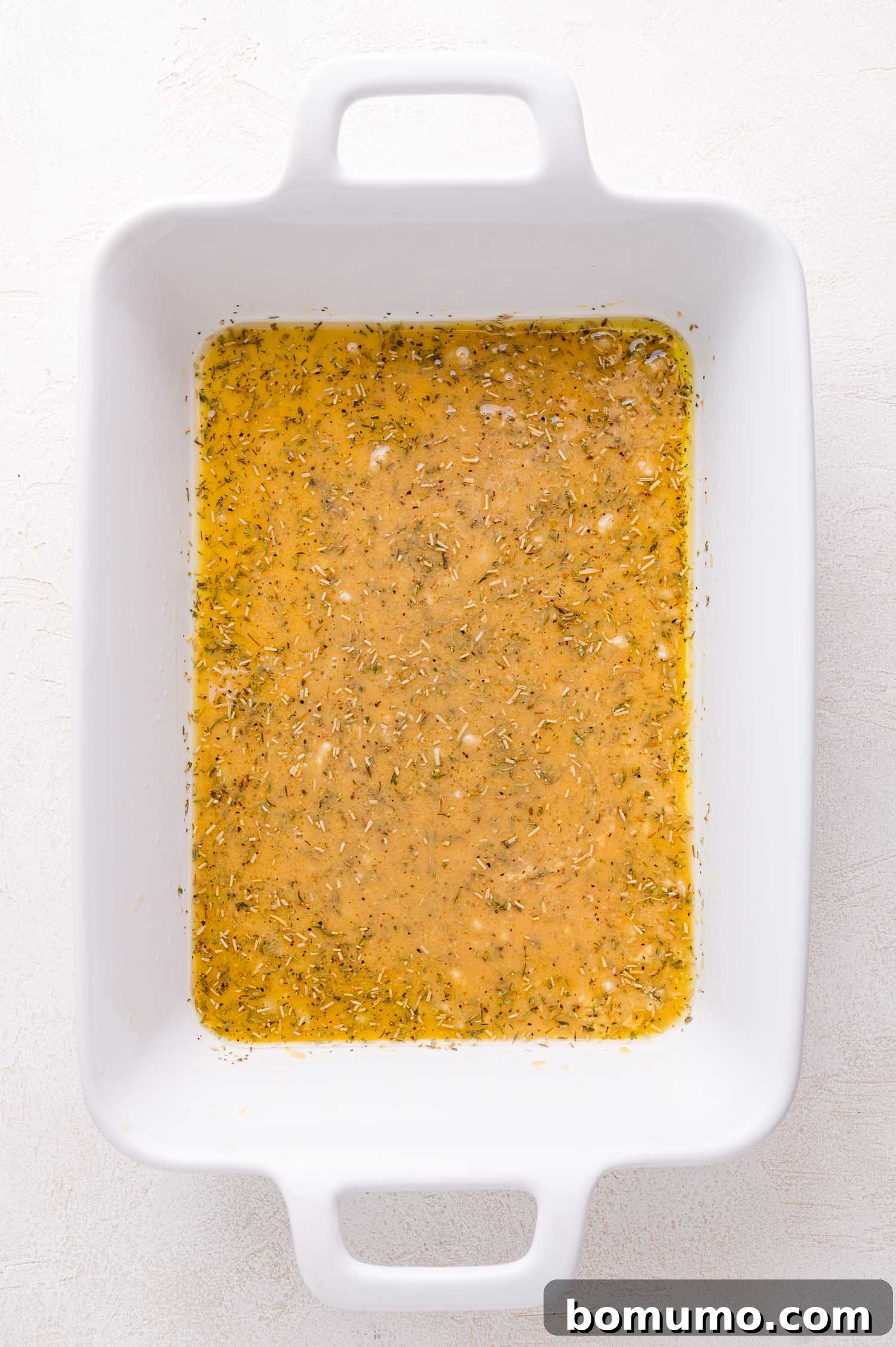 White rectangular baking dish filled with a yellowish marinade, herbs, and spices, freshly mixed on a light-colored surface.