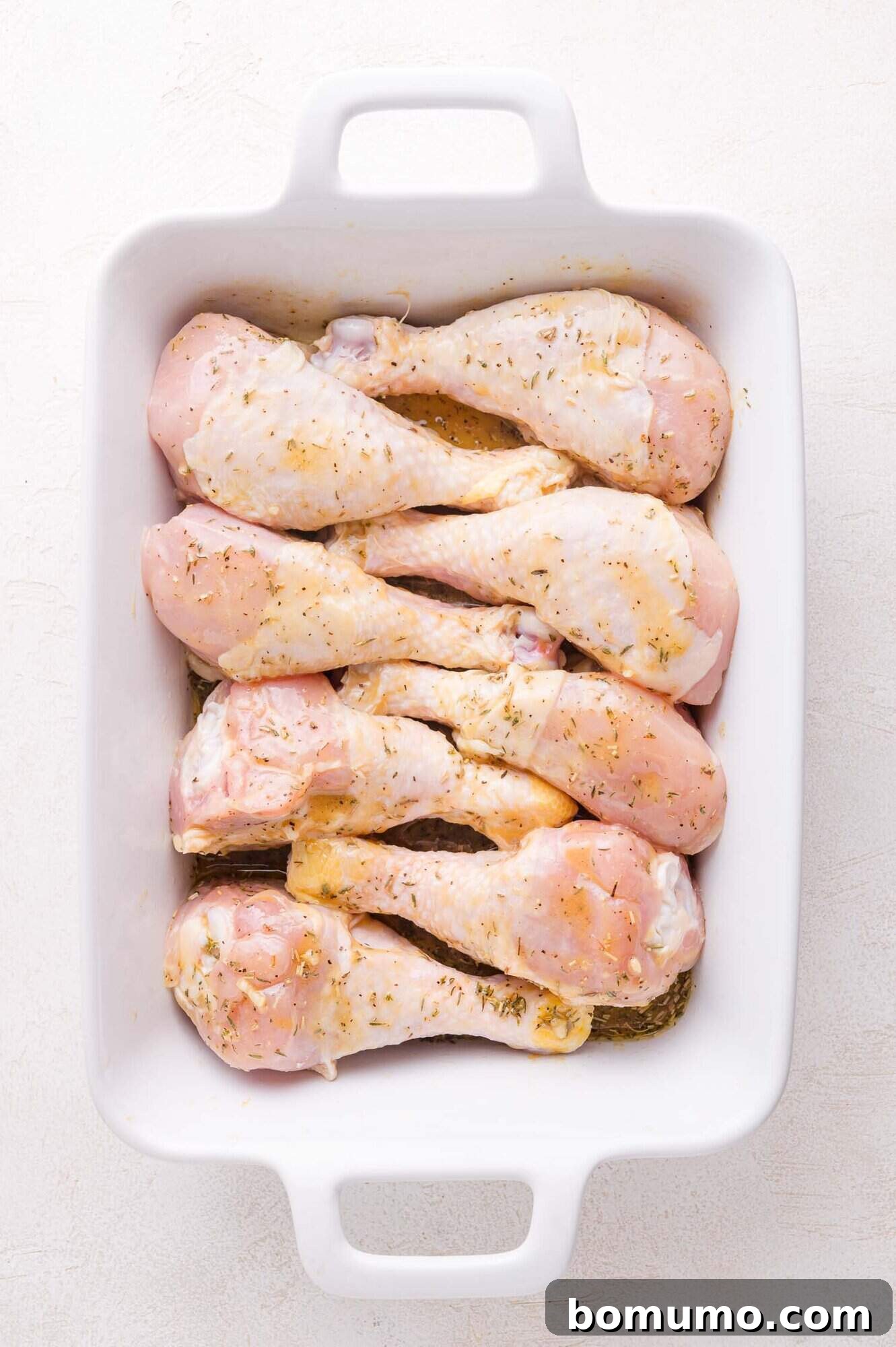Raw, seasoned chicken drumsticks marinating in a single layer inside a white rectangular baking dish on a light surface, fully coated.