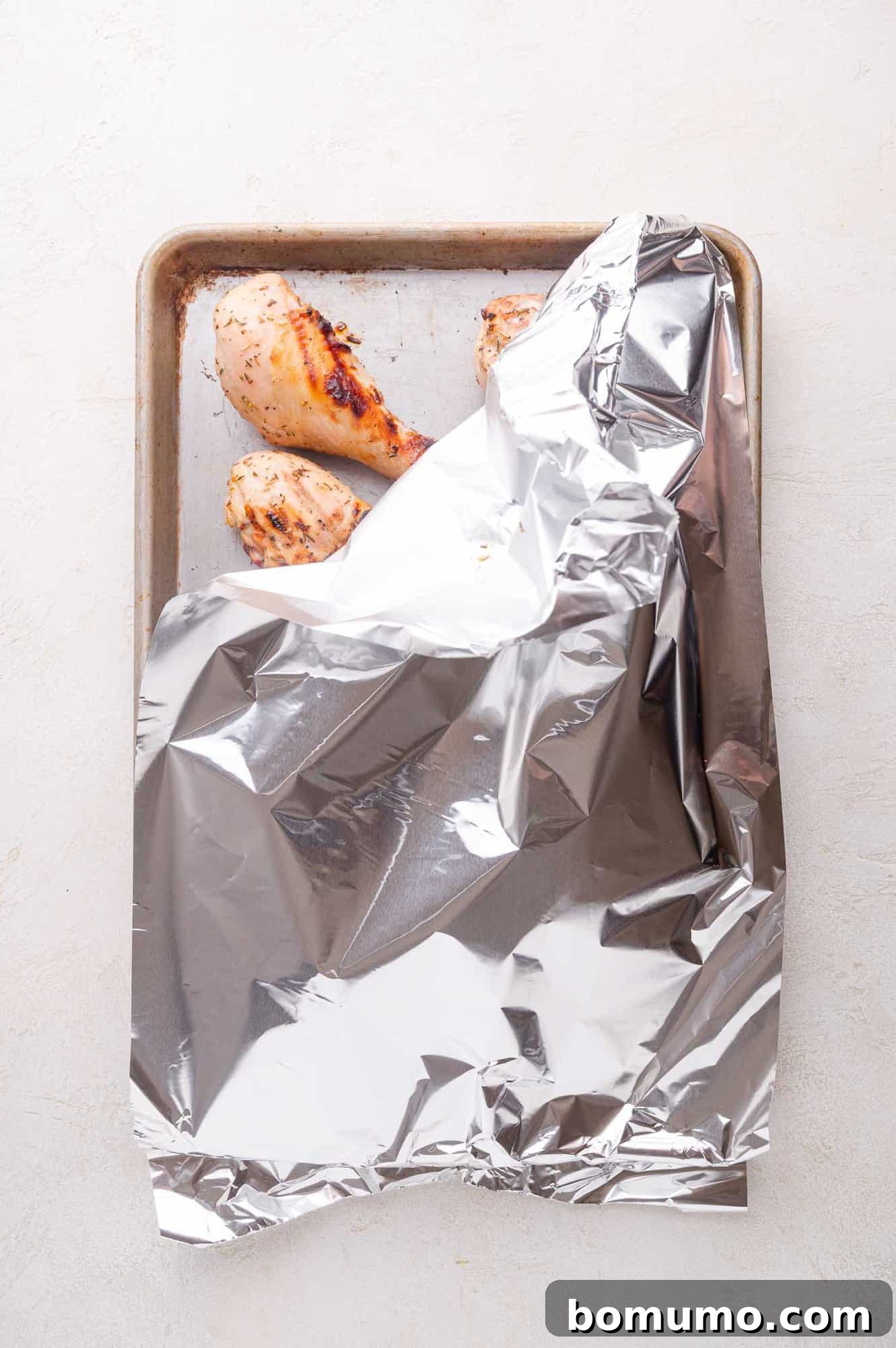 A baking sheet with cooked chicken drumsticks partially covered by a sheet of aluminum foil, resting on a light surface.