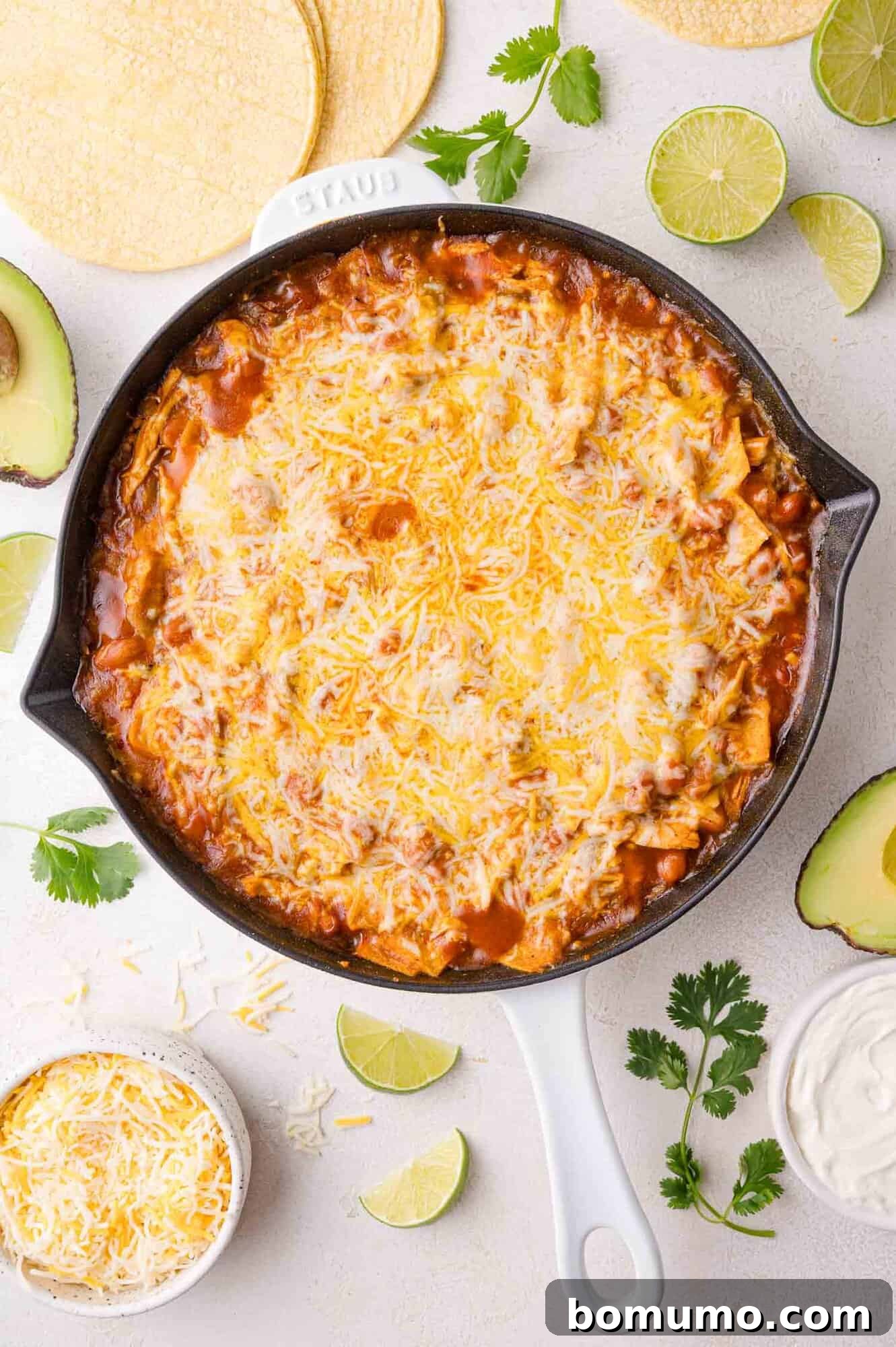 Quick Skillet Chicken Enchilada 15 A perfectly baked skillet filled with a cheesy chicken enchilada casserole sits on a clean white surface, surrounded by fresh avocado halves, bright lime wedges, fragrant cilantro, extra shredded cheese, and a stack of corn tortillas, creating a vibrant culinary scene.