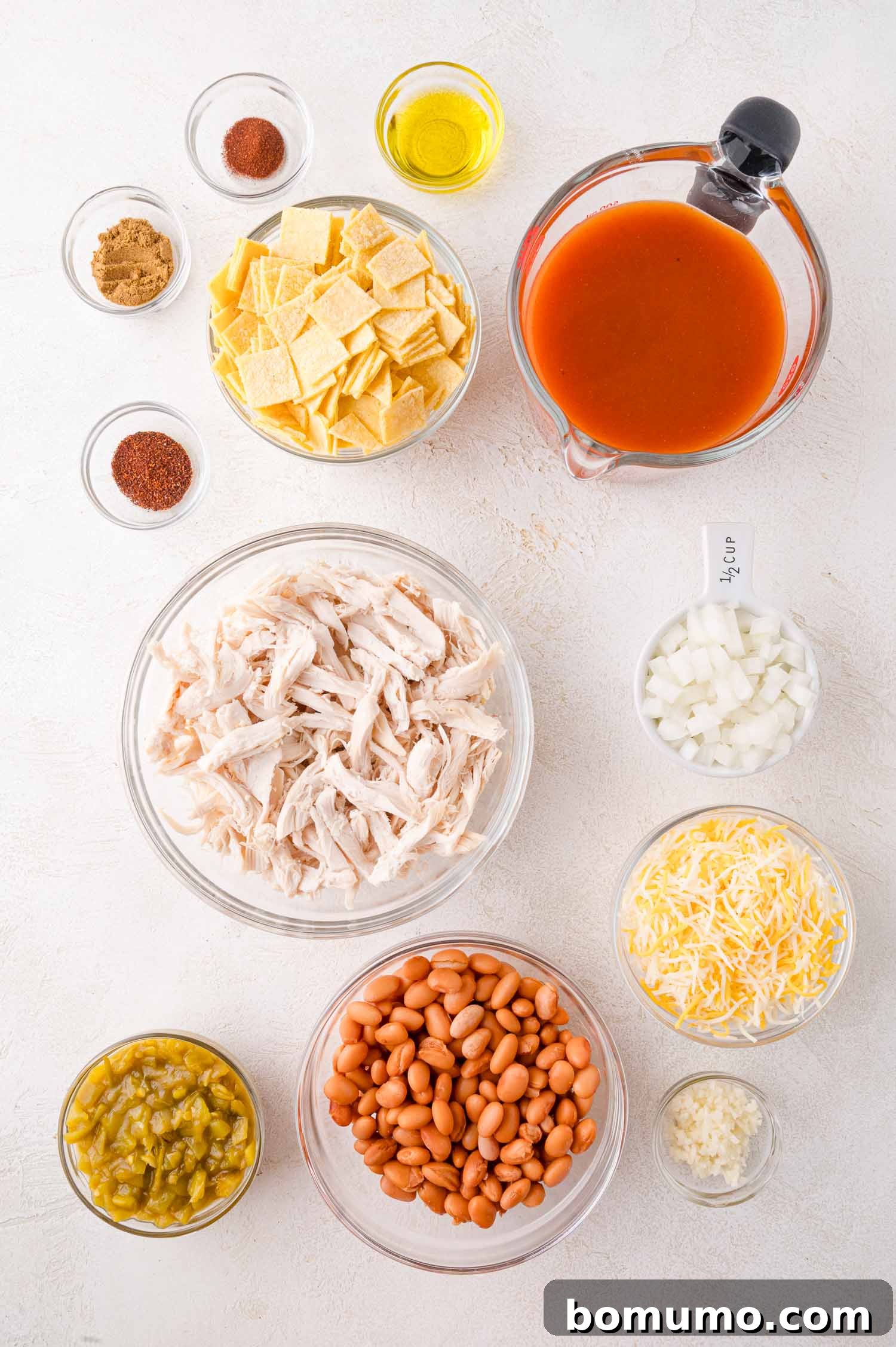 Quick Skillet Chicken Enchilada 4 Various essential ingredients for chicken enchilada skillet are neatly arranged in separate bowls on a white surface, including shredded chicken, pinto beans, shredded cheese, chopped onions, red enchilada sauce, cut corn tortilla pieces, and a selection of seasonings.