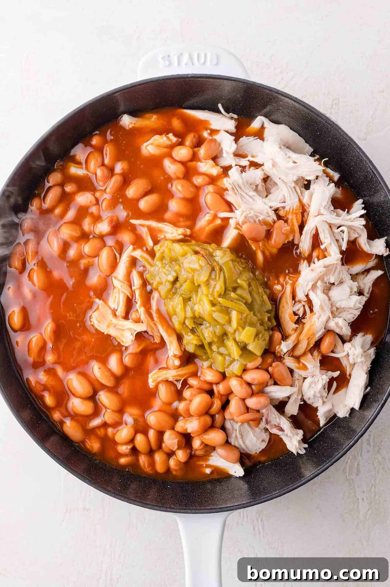Quick Skillet Chicken Enchilada 8 A skillet containing pinto beans in a rich red sauce, along with shredded chicken and green chiles, all partially mixed.