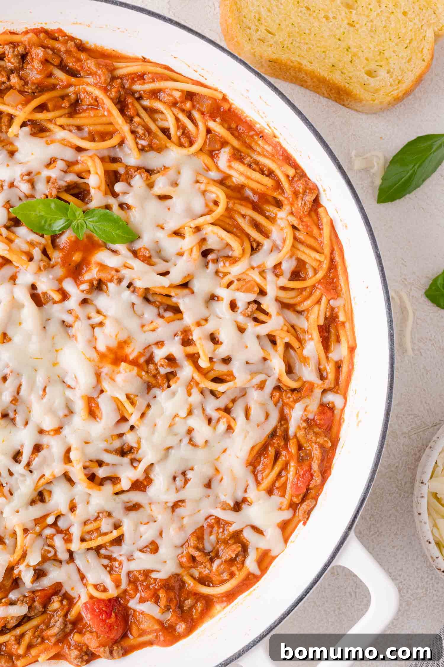 Delicious one-pan spaghetti with meat sauce, topped with bubbling melted mozzarella cheese, garnished with fresh basil leaves. A side of golden garlic bread completes the comforting meal.