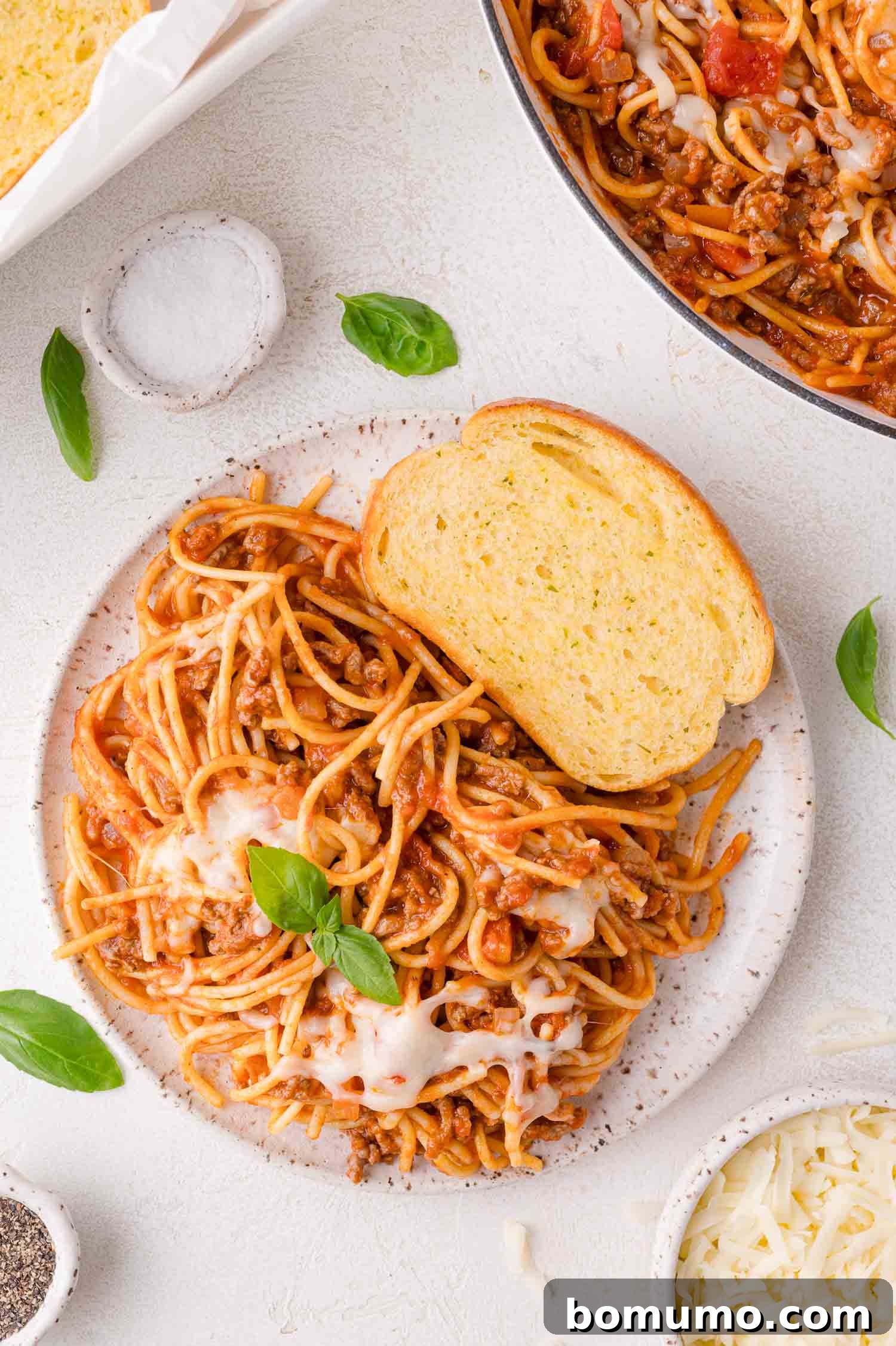 A beautifully arranged plate featuring a generous serving of spaghetti with rich meat sauce and melted cheese, elegantly garnished with fresh basil leaves. Two golden slices of garlic bread rest alongside on a pristine white surface, completing a classic and comforting meal presentation.