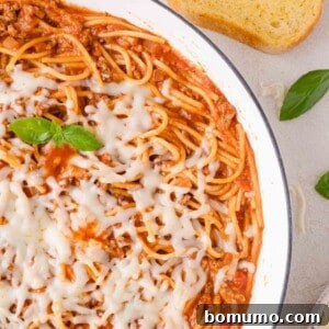 A skillet of one pan spaghetti topped with melted cheese, garnished with basil, sits on a light surface with a slice of bread nearby.