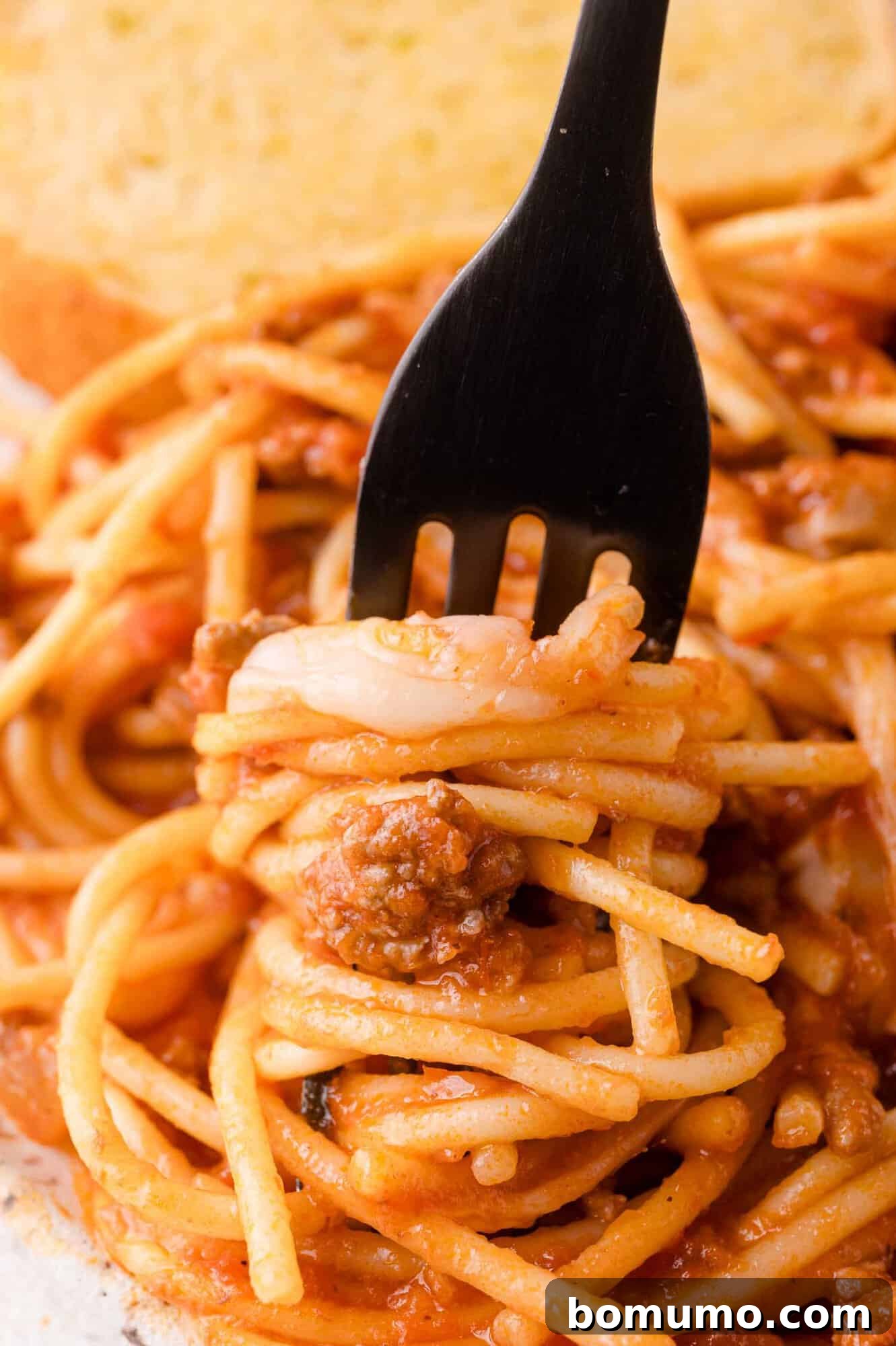 A black fork gracefully twirls a generous portion of spaghetti with meat sauce, ready to be enjoyed. A crisp piece of garlic bread is visible in the background, adding to the allure of this comforting dish.