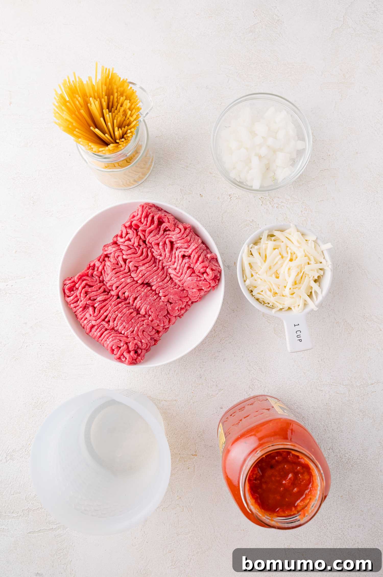A vibrant flat lay of key ingredients for one-pan spaghetti: uncooked spaghetti noodles, finely chopped onions, a mound of shredded mozzarella cheese, raw ground beef, a jar of rich marinara sauce, and a clear measuring cup filled with water, all artfully arranged on a clean white surface.