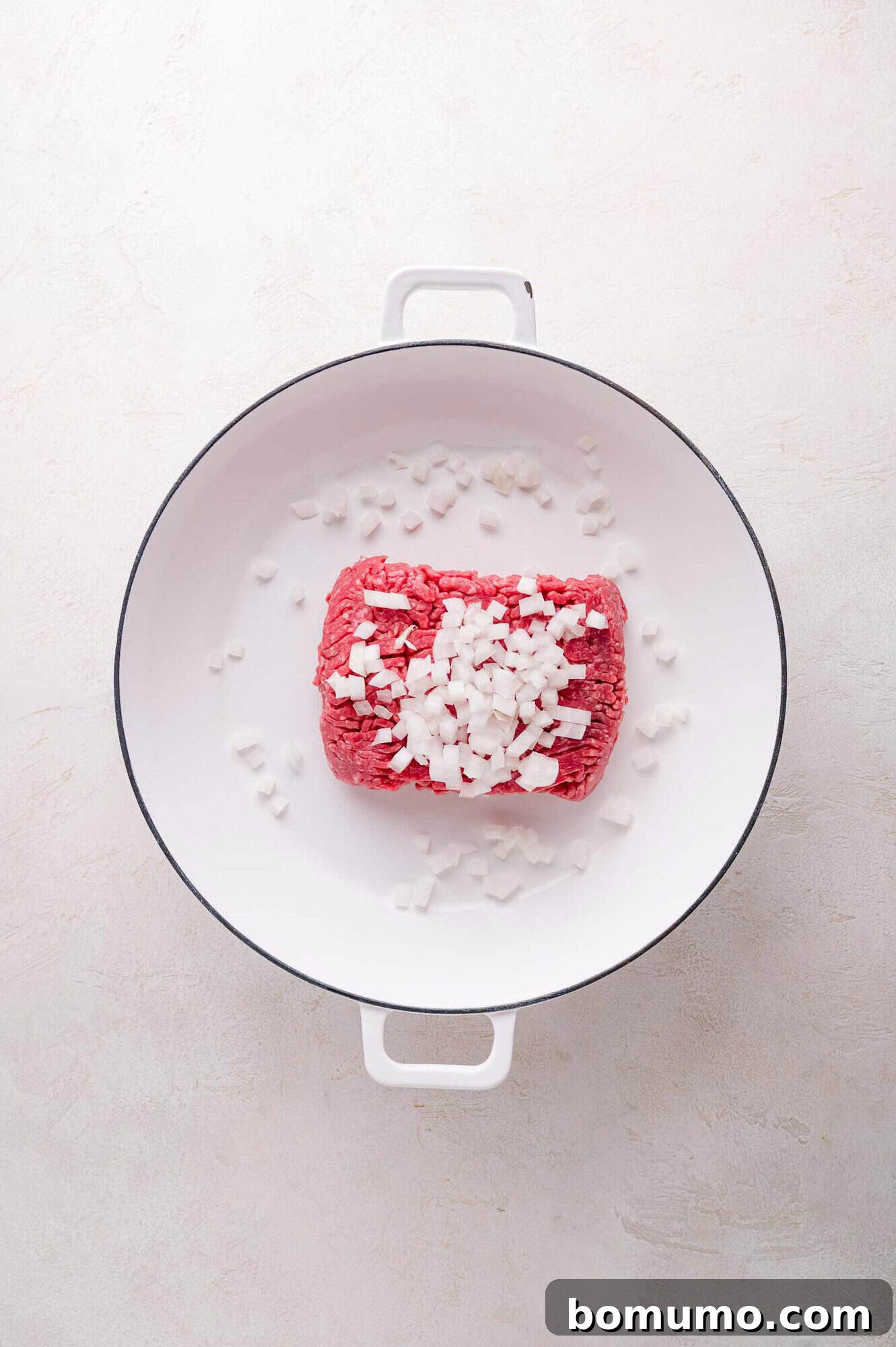 A block of raw ground beef and chopped yellow onions placed side-by-side in a large white pot, ready for cooking on a light-colored kitchen surface.