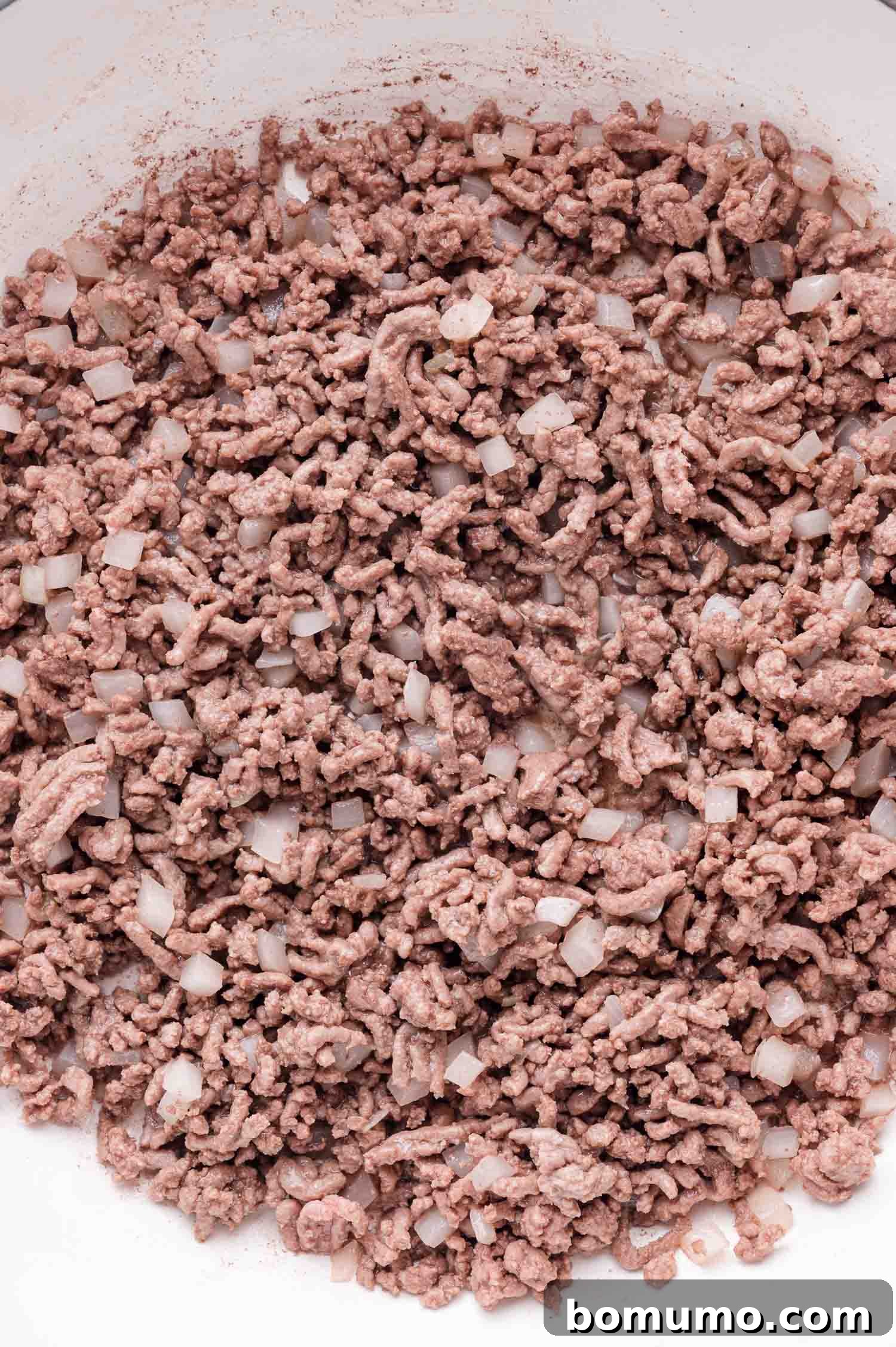 Close-up view of ground beef and chopped onions sizzling and browning together in a hot pan, being stirred with a black spatula.
