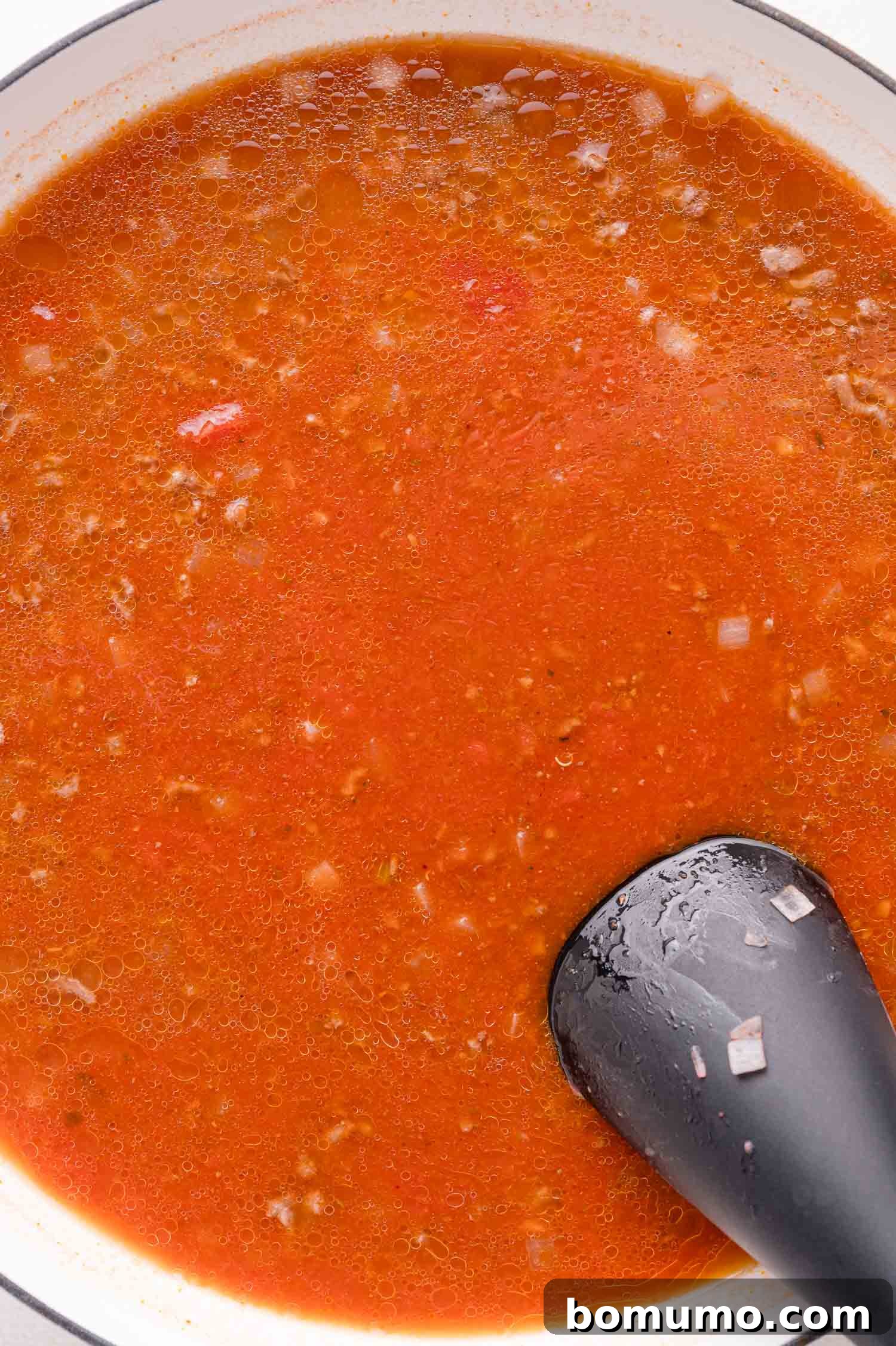 A delicious pot of tomato-based sauce, featuring visible ground meat and diced onions, is actively being stirred with a sleek black kitchen utensil, creating a harmonious blend of flavors.