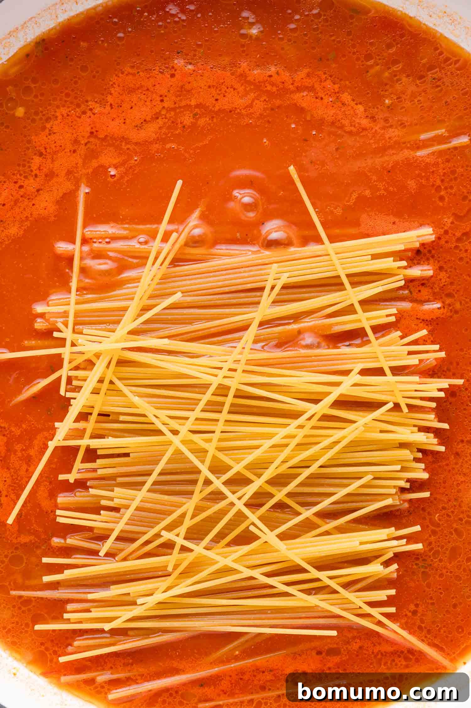 Uncooked spaghetti noodles carefully placed into a bubbling pot of rich red tomato sauce, beginning to soften and absorb the flavors.