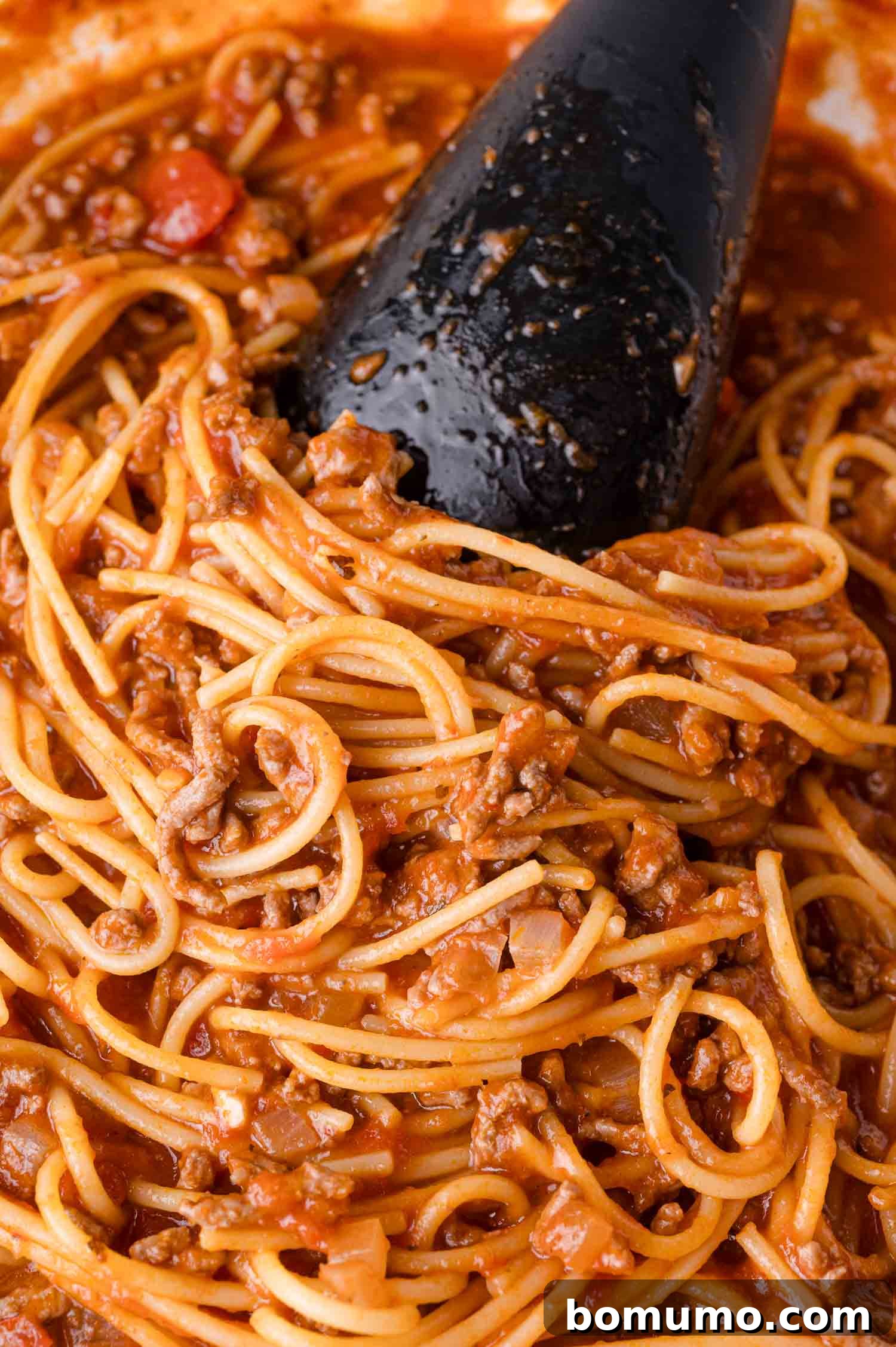 A close-up view of cooked spaghetti perfectly mixed with ground meat and a rich tomato sauce, ready to be served. A black serving spoon is partially visible, hinting at the deliciousness within.