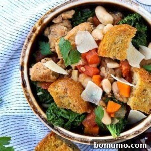 Overhead of earthenware bowl containing sausage, bean, and kale soup garnished with large croutons.