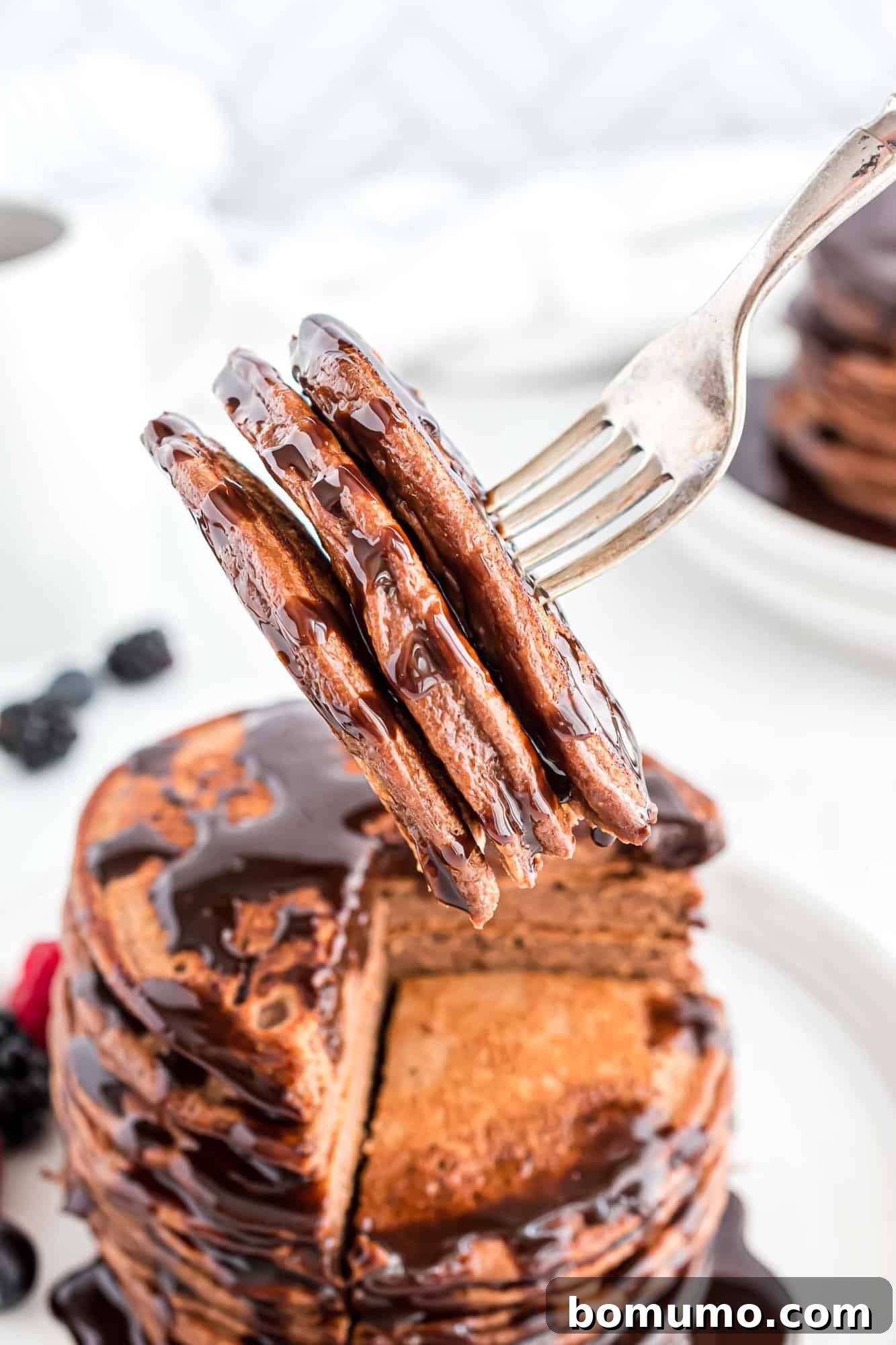 A close-up of a chocolate protein pancake on a fork, drizzled with rich chocolate syrup.