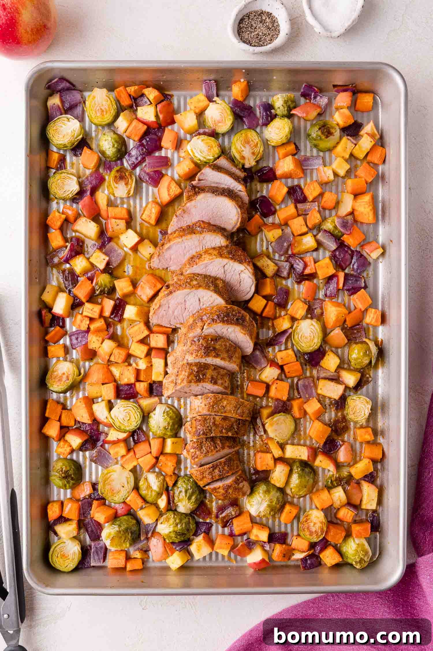 A baking sheet with sliced pork tenderloin surrounded by roasted Brussels sprouts, sweet potatoes, apples, and red onions. Salt and pepper bowls are nearby.