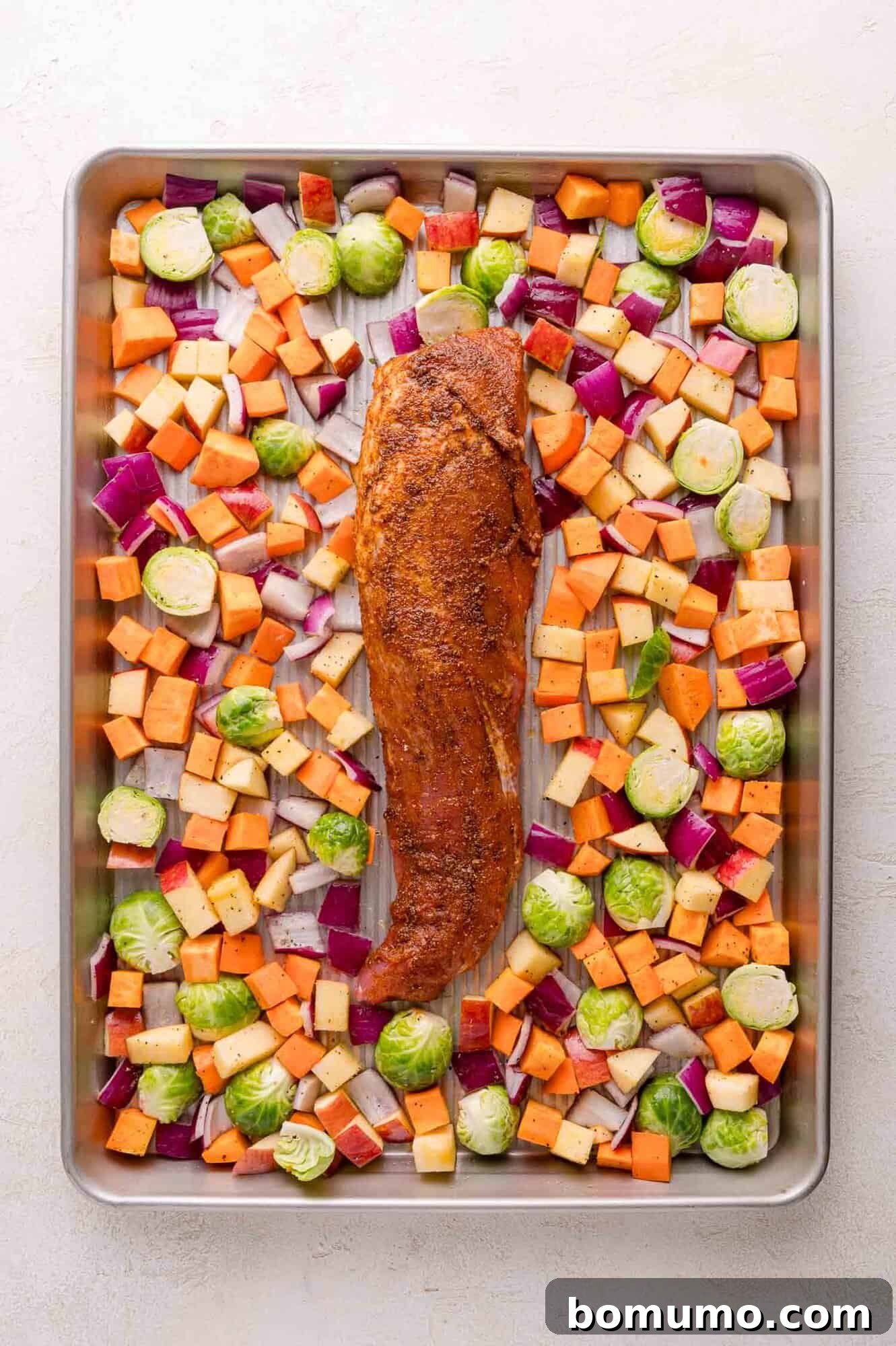 A sheet pan with a seasoned pork tenderloin in the center, surrounded by diced sweet potatoes, Brussels sprouts, red onions, and apples.
