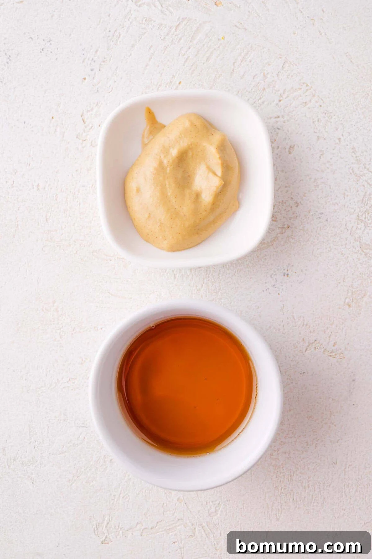 A small white bowl of creamy dijon mustard above a small white bowl of maple syrup, both on a light textured surface.