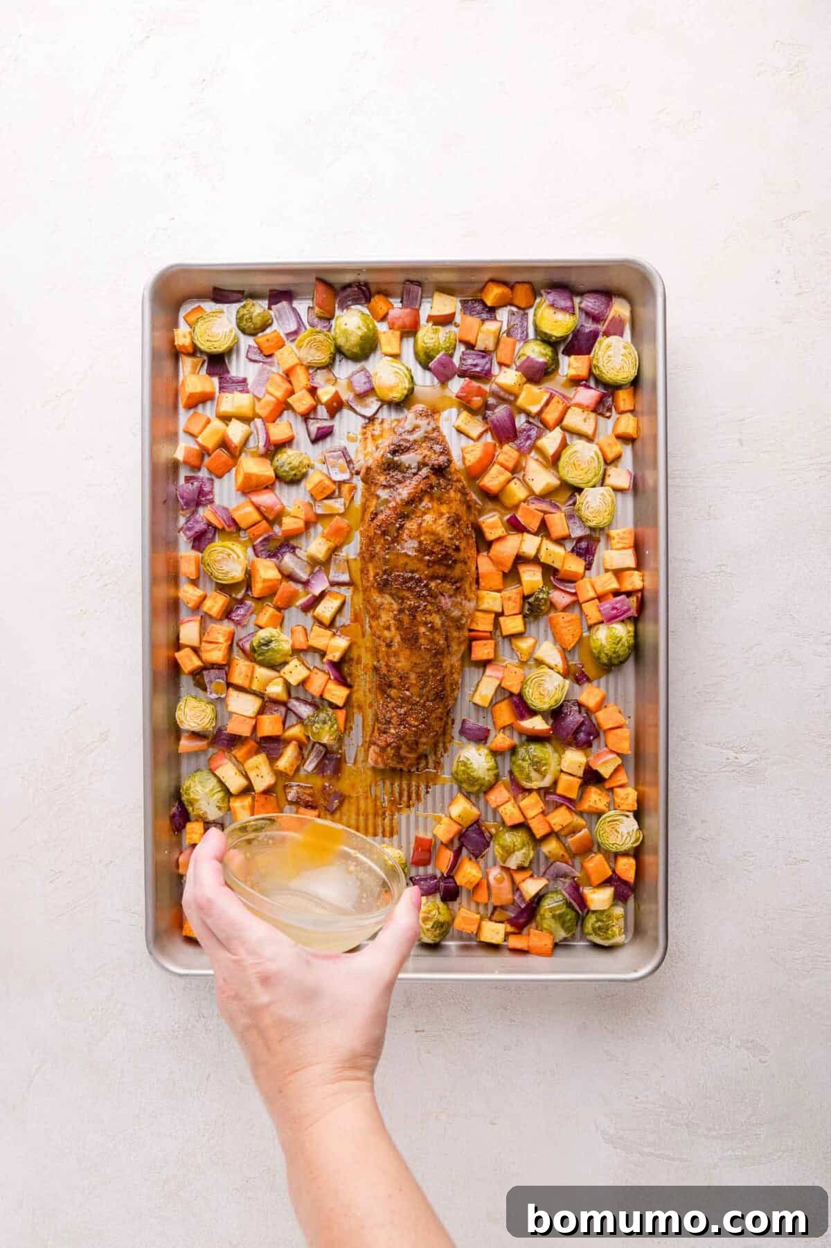 A hand holds a bowl of maple syrup and mustard mixture above a baking sheet with roasted pork tenderloin, sweet potatoes, Brussels sprouts, and red onions.