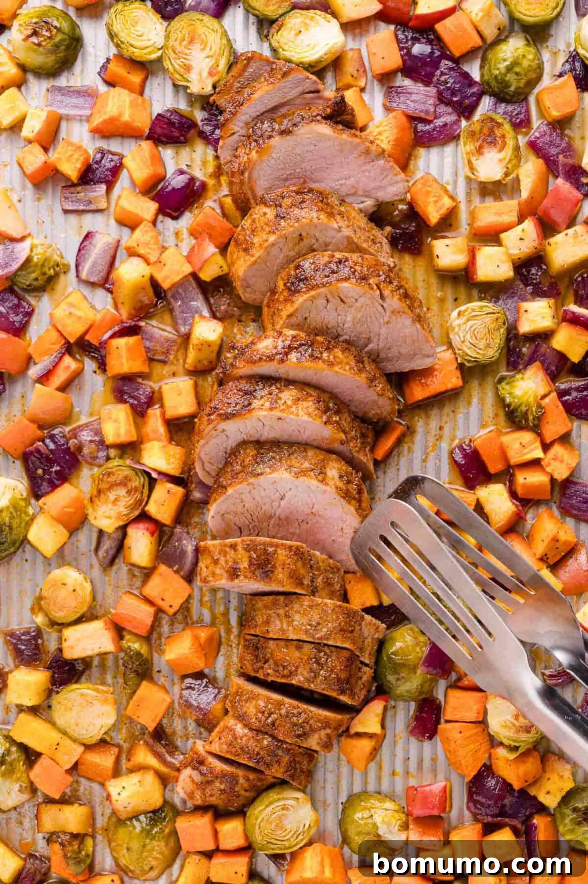Sliced pork tenderloin on a baking sheet surrounded by roasted sweet potatoes, Brussels sprouts, and red onions, with metal tongs beside the pork.