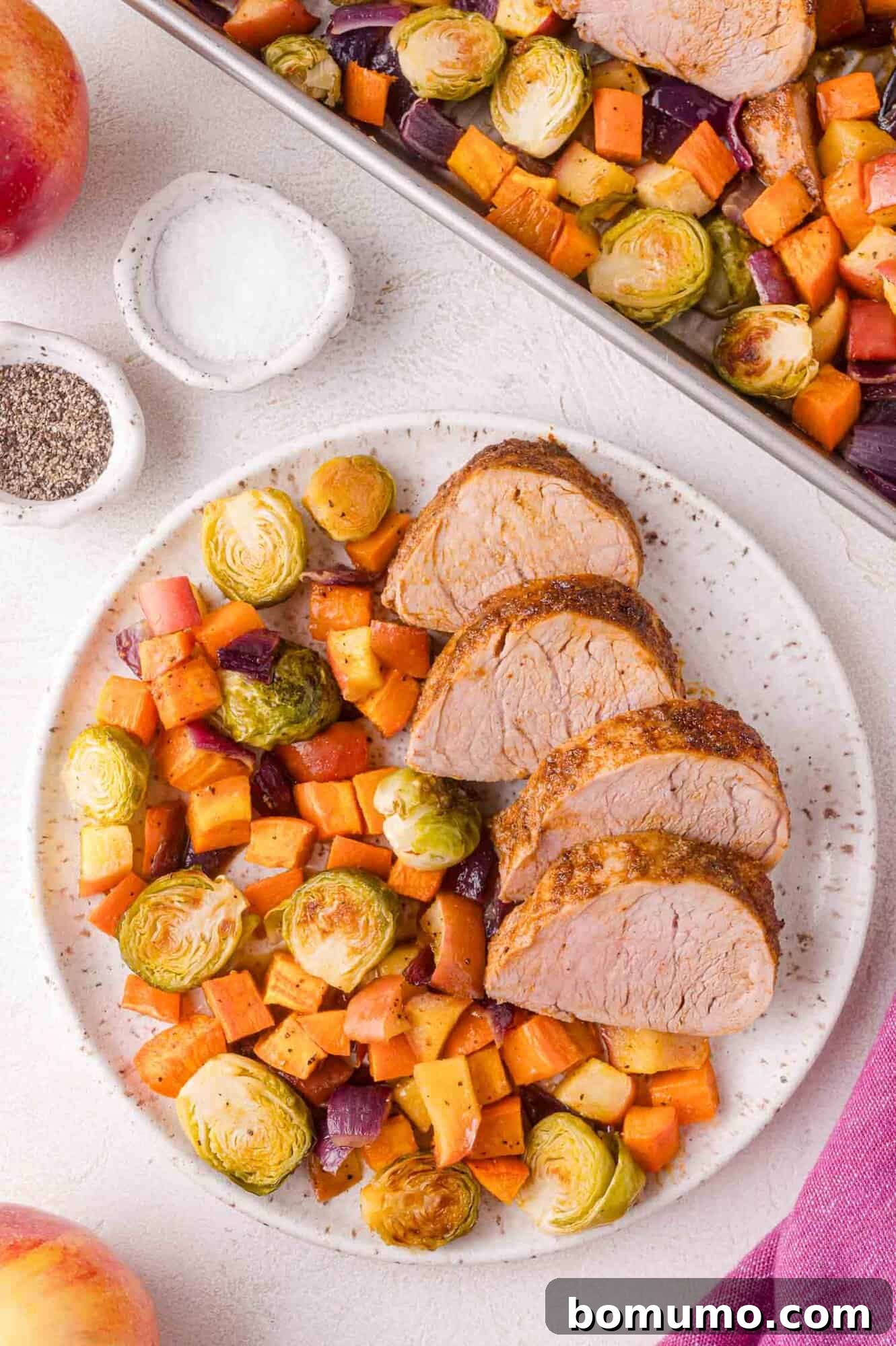 A plate with three slices of cooked pork tenderloin served alongside roasted Brussels sprouts, sweet potatoes, onions, and apples. A baking sheet with more food is in the background.
