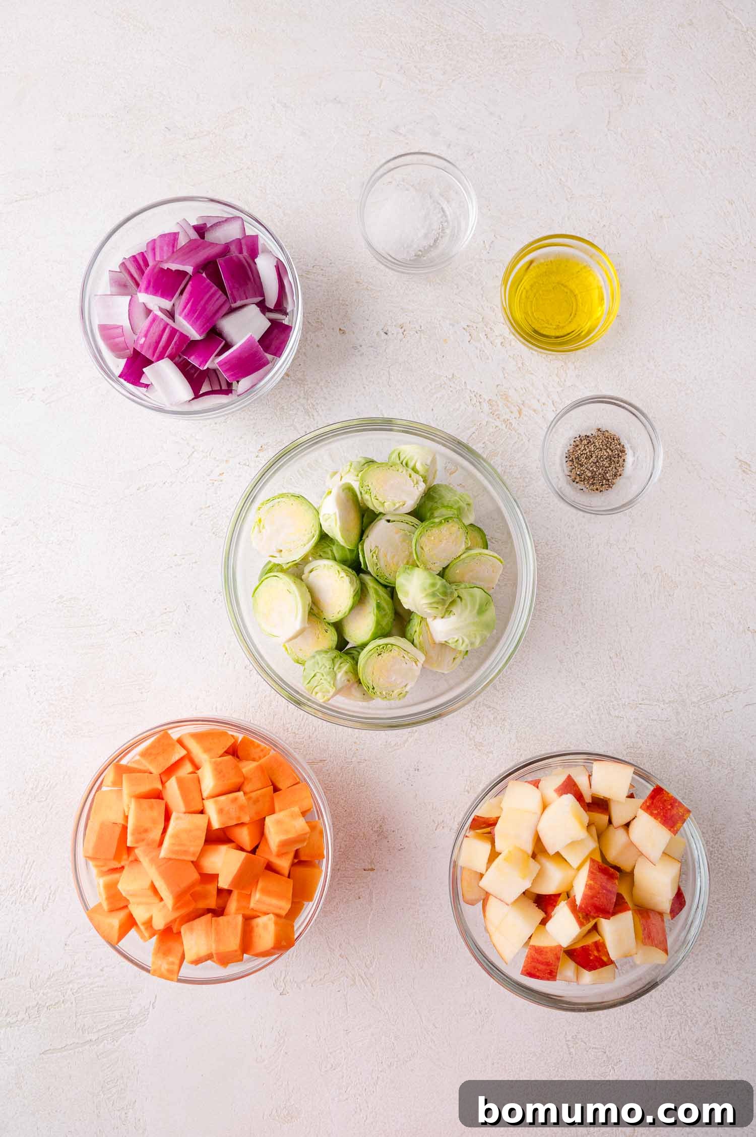 Five glass bowls with chopped red onion, halved Brussels sprouts, diced sweet potato, and diced apples, plus small bowls of oil, salt, and pepper on a light surface.