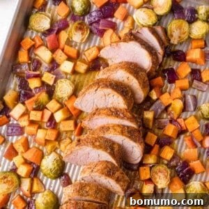 Sliced pork tenderloin with roasted Brussels sprouts, red onions, sweet potatoes, and root vegetables on a baking sheet.