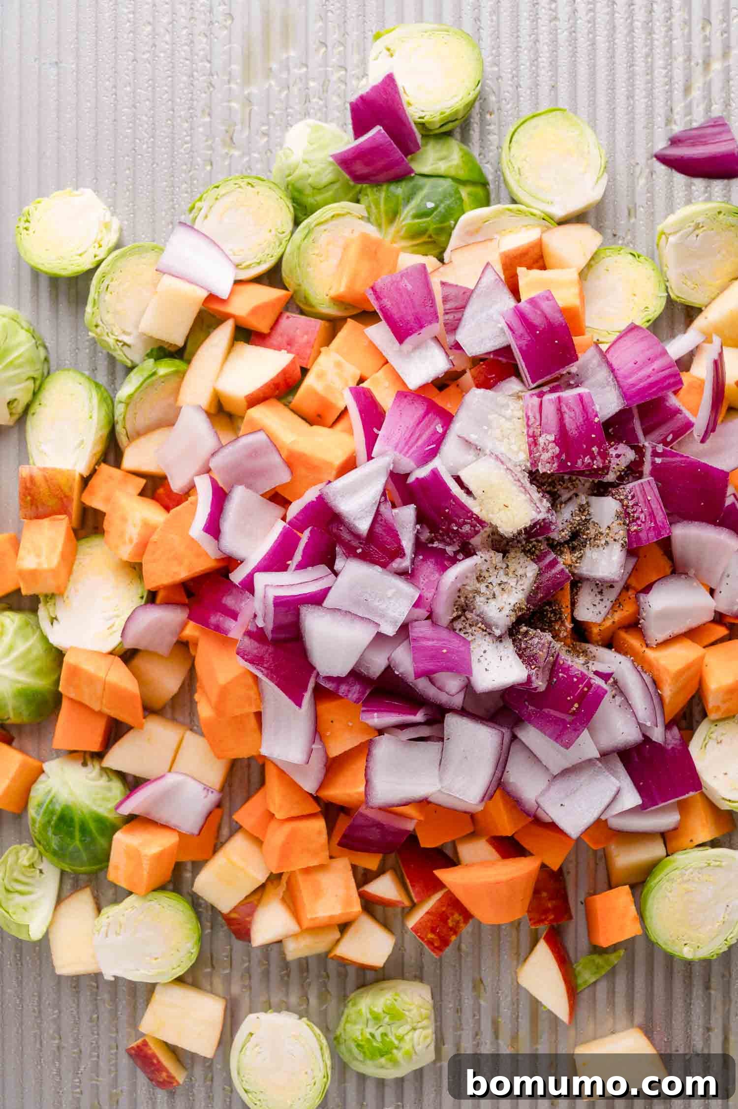Chopped Brussels sprouts, red onion, sweet potato, and apples, on a baking sheet, sprinkled with salt and pepper.