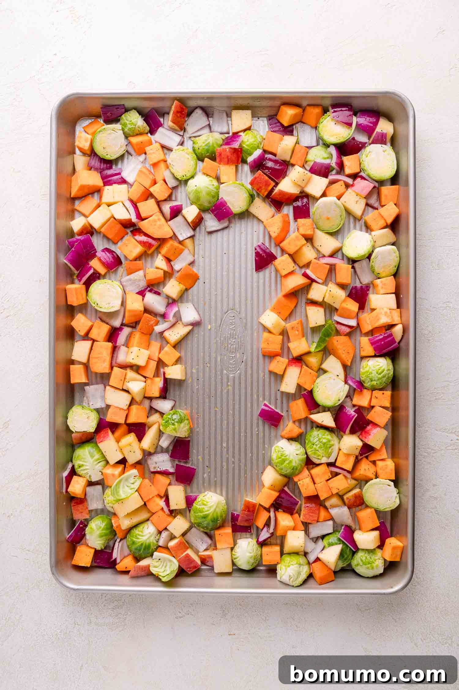 Chopped sweet potatoes, Brussels sprouts, red onions, and aples spread on a metal baking sheet, ready for roasting.