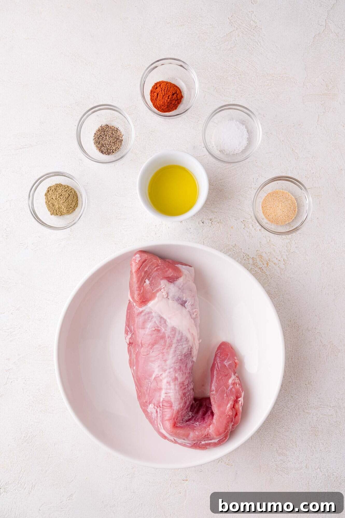 Raw pork tenderloin in a white bowl surrounded by bowls of olive oil, salt, pepper, paprika, garlic powder, and dried herbs on a light surface.