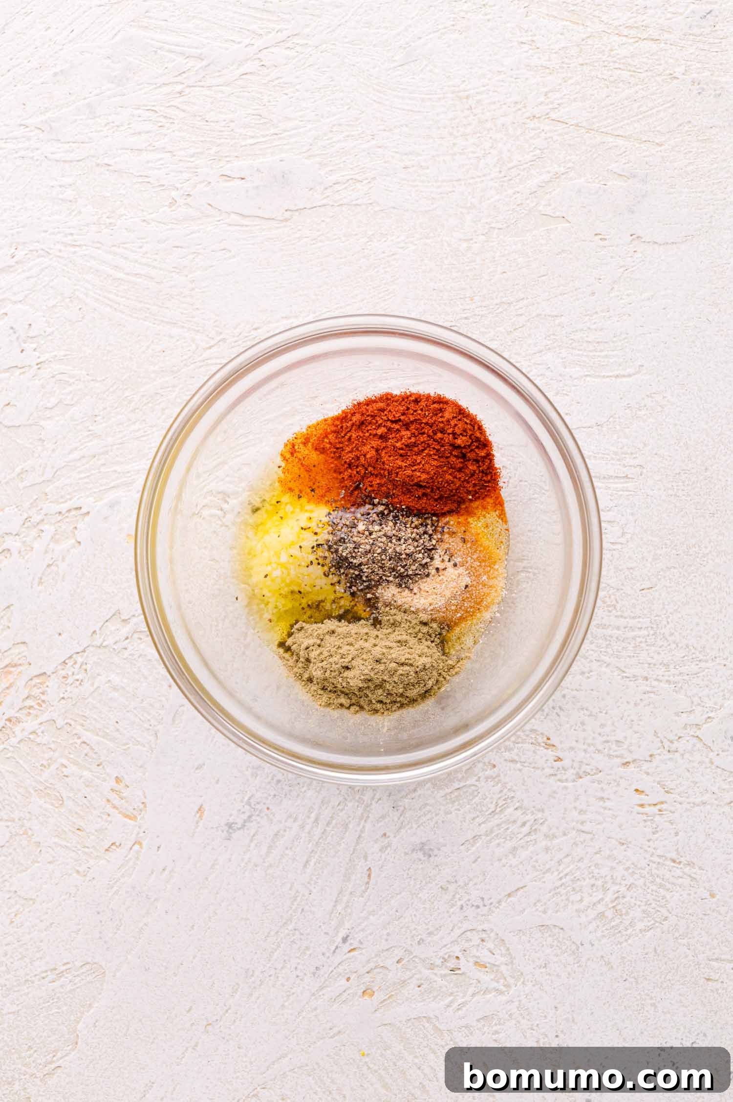 A glass bowl containing ground spices, arranged in separate piles on a textured white surface.