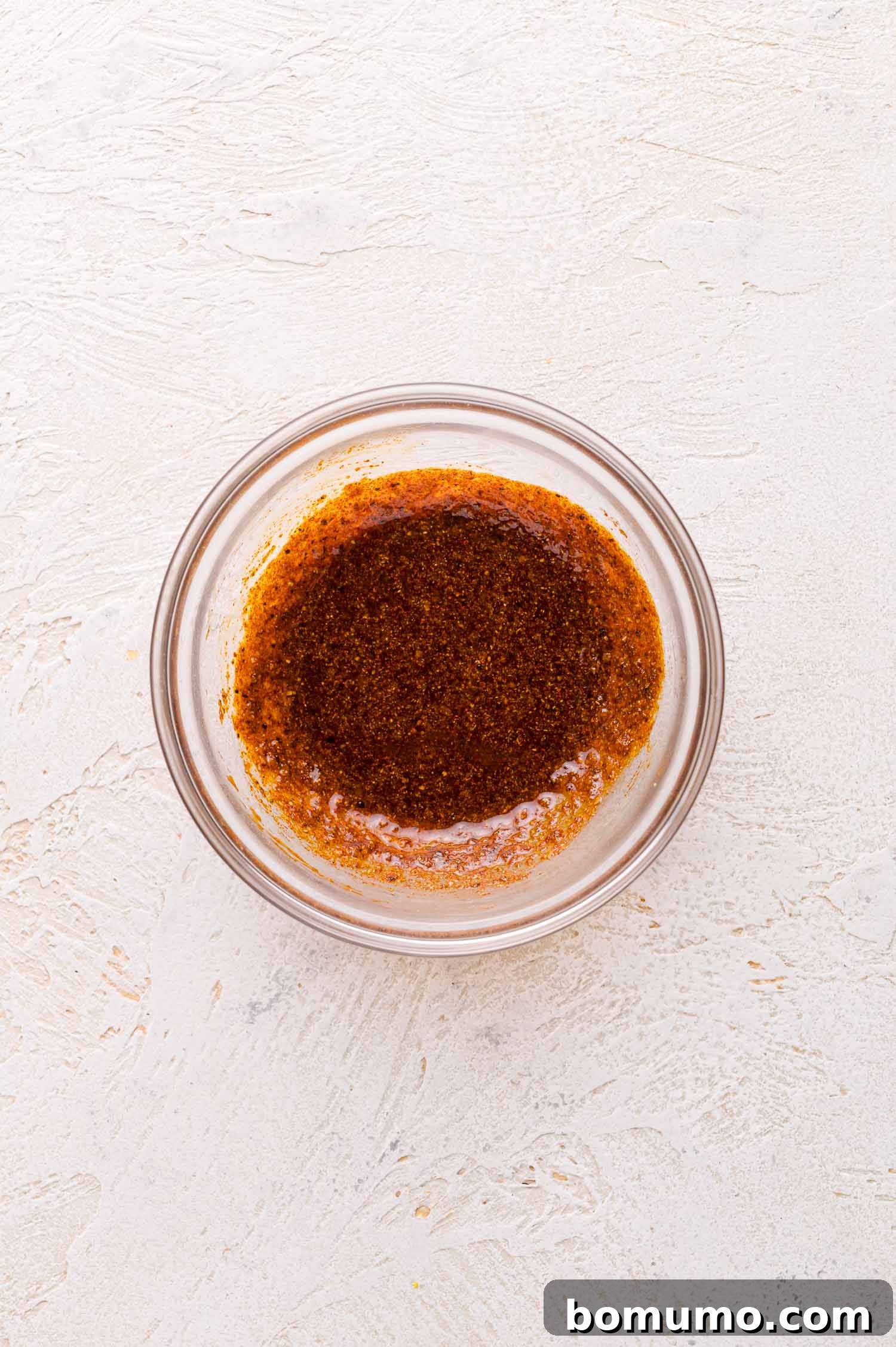 A small glass bowl filled with a dark brown spice mixture on a textured light-colored surface.
