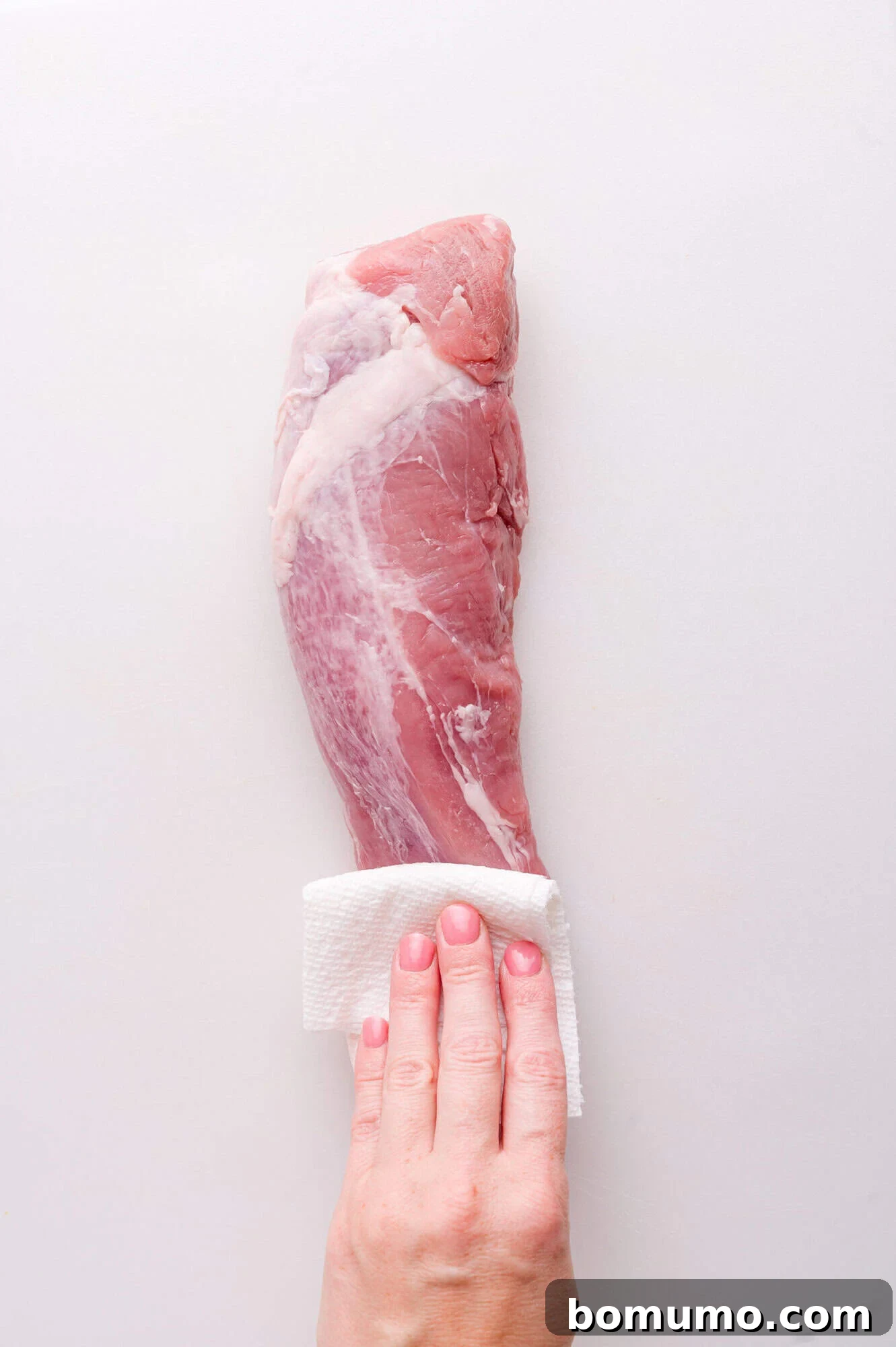 A hand uses a paper towel to pat dry a raw pork tenderloin on a white surface.