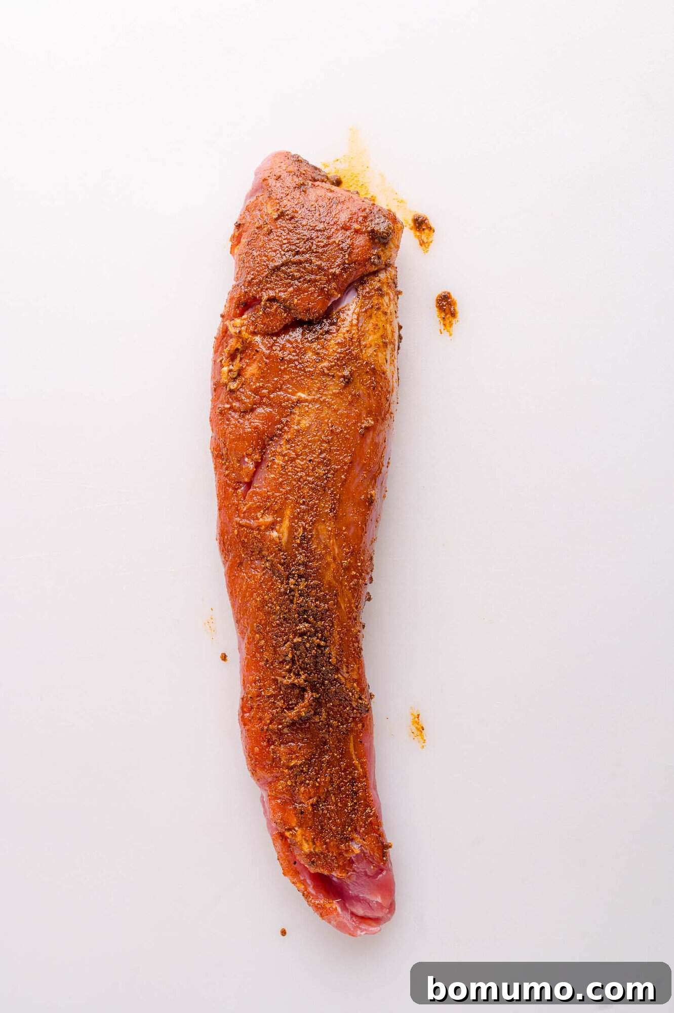 A raw pork tenderloin coated with a reddish-brown spice rub sits on a white surface.