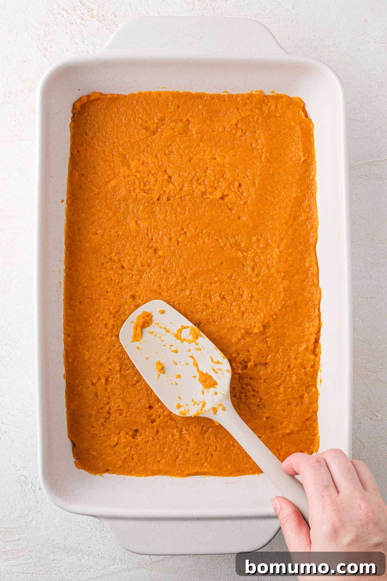 A spatula smoothing the sweet potato mixture into an oven-safe white casserole dish, preparing it for baking and ensuring an even layer.