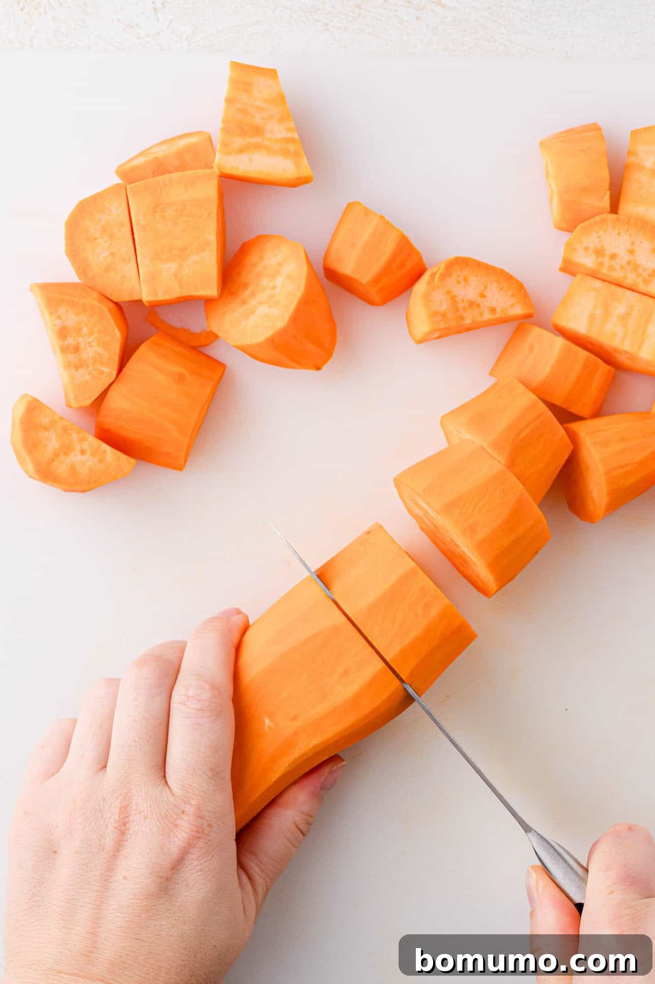 A sharp knife slicing a sweet potato into uniform chunks on a cutting board, preparing them for cooking and ensuring even tenderness.