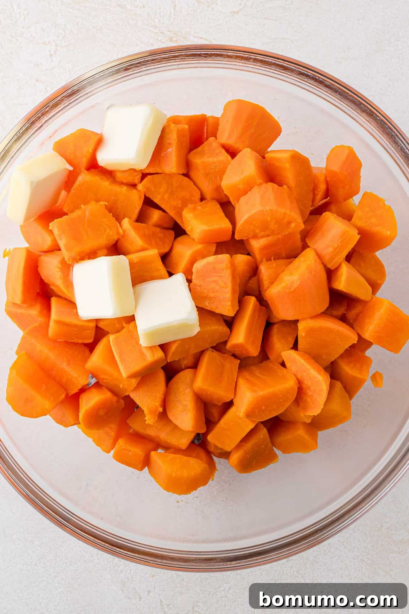 Cooked sweet potato chunks and small pieces of butter placed together in a large glass mixing bowl, poised for mashing and blending.