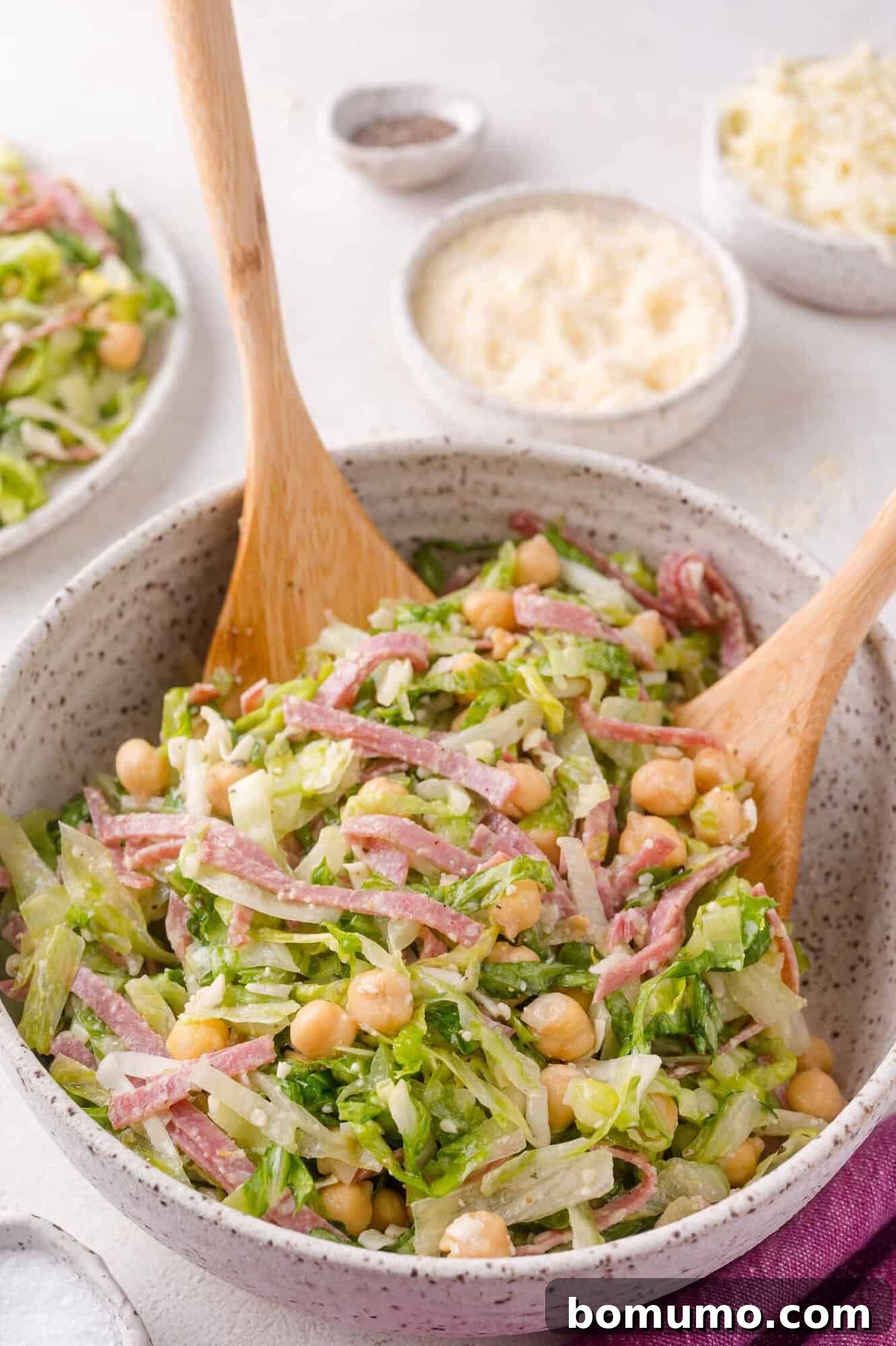 A vibrant bowl of chopped salad, showcasing fresh lettuce, golden chickpeas, thinly sliced salami, and grated cheese, accompanied by two elegant wooden salad servers. In the background, smaller dishes of cheese and black pepper add to the inviting scene.