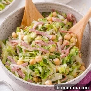 A bowl of La Scala chopped salad with lettuce, chickpeas, sliced salami, and cheese, mixed and served with two wooden salad utensils.