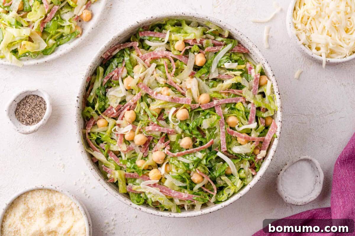 A beautifully presented bowl of La Scala chopped salad, featuring finely shredded lettuce, tender chickpeas, savory salami strips, and generous portions of shredded cheese, all resting on a clean, light surface. The scene is further enhanced by an arrangement of small bowls filled with fresh ingredients, highlighting the salad's vibrant components.