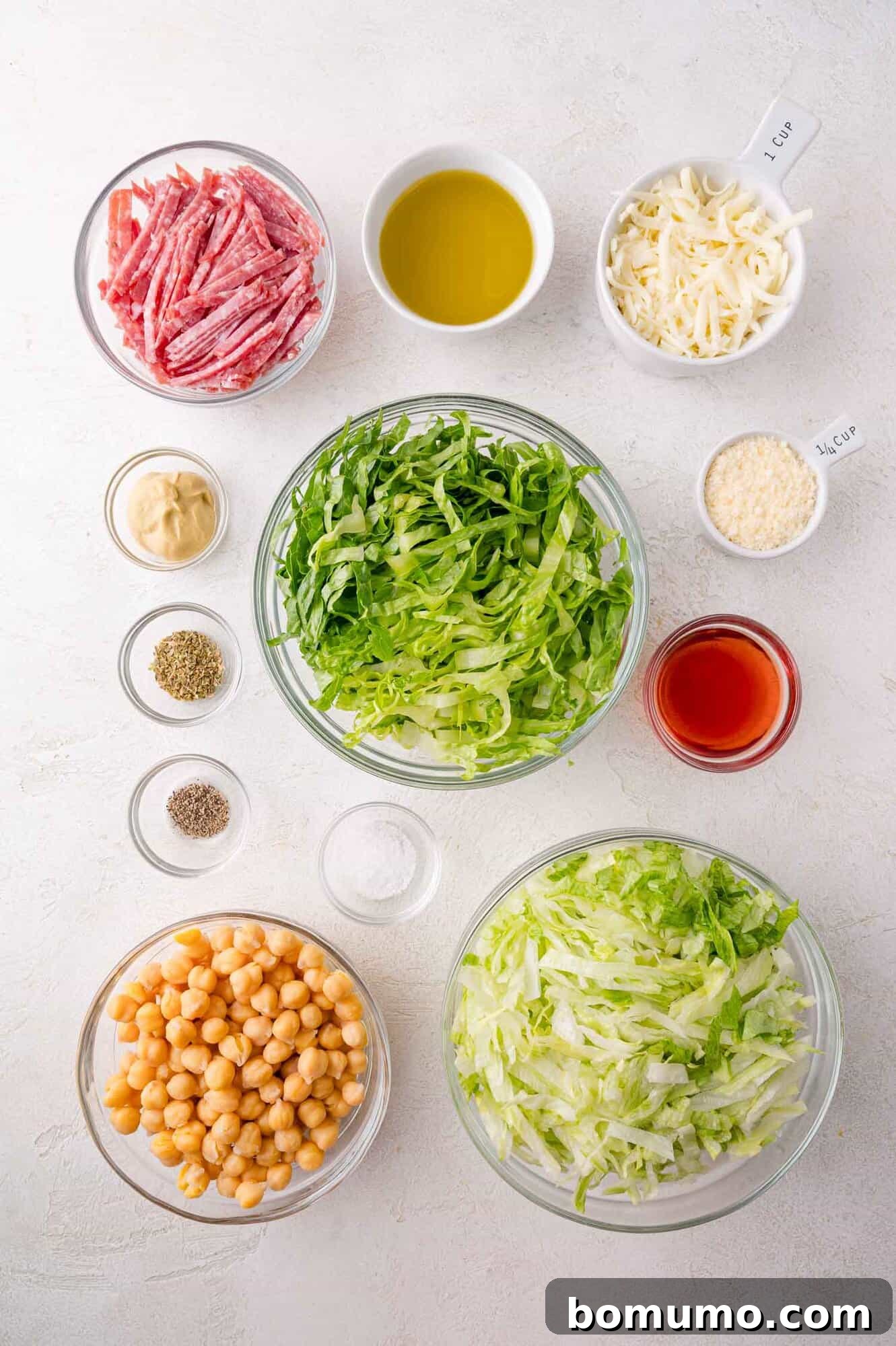 Bowls neatly arranged with the fresh components for La Scala chopped salad: shredded lettuce, cooked chickpeas, thinly sliced salami, a mix of shredded and grated cheeses, alongside bottles of olive oil, vinegar, and small dishes of mustard and various seasonings, all set on a pristine white surface.