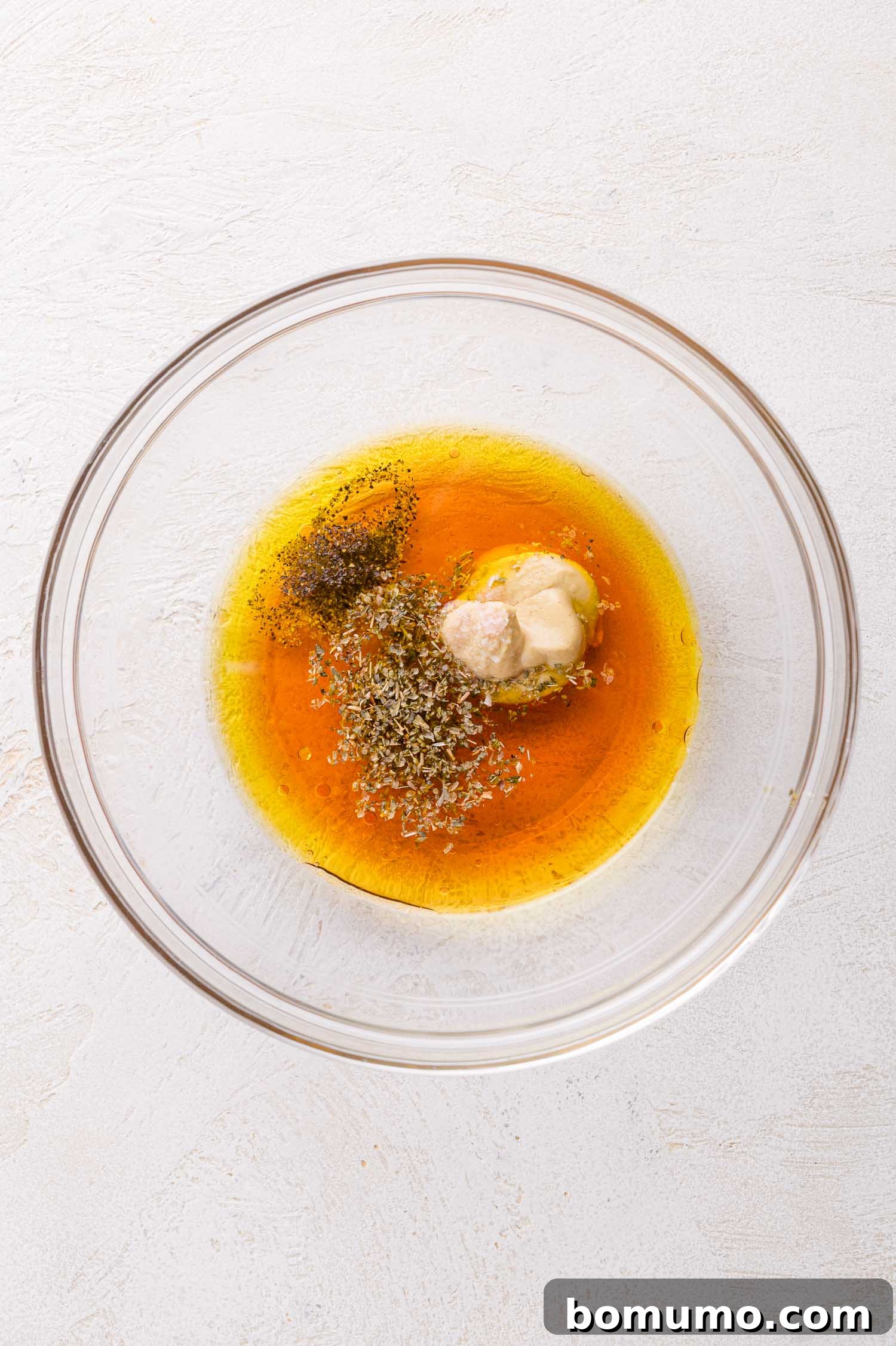 A clear glass bowl holding the initial liquid ingredients for the dressing: olive oil, a dollop of mustard, and dried herbs, all perfectly positioned on a clean white surface, awaiting emulsification.