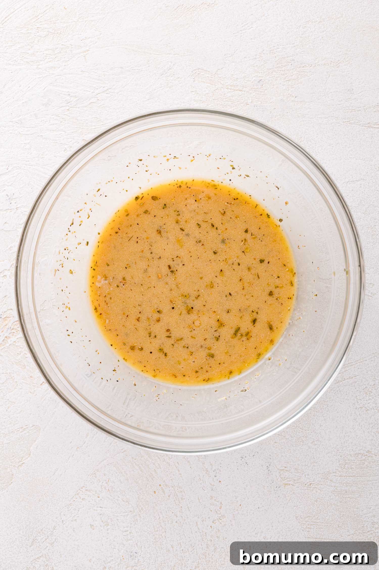A transparent glass bowl displaying a light brown, creamy dressing, beautifully emulsified and ready to infuse flavor, resting elegantly on a pristine white surface.